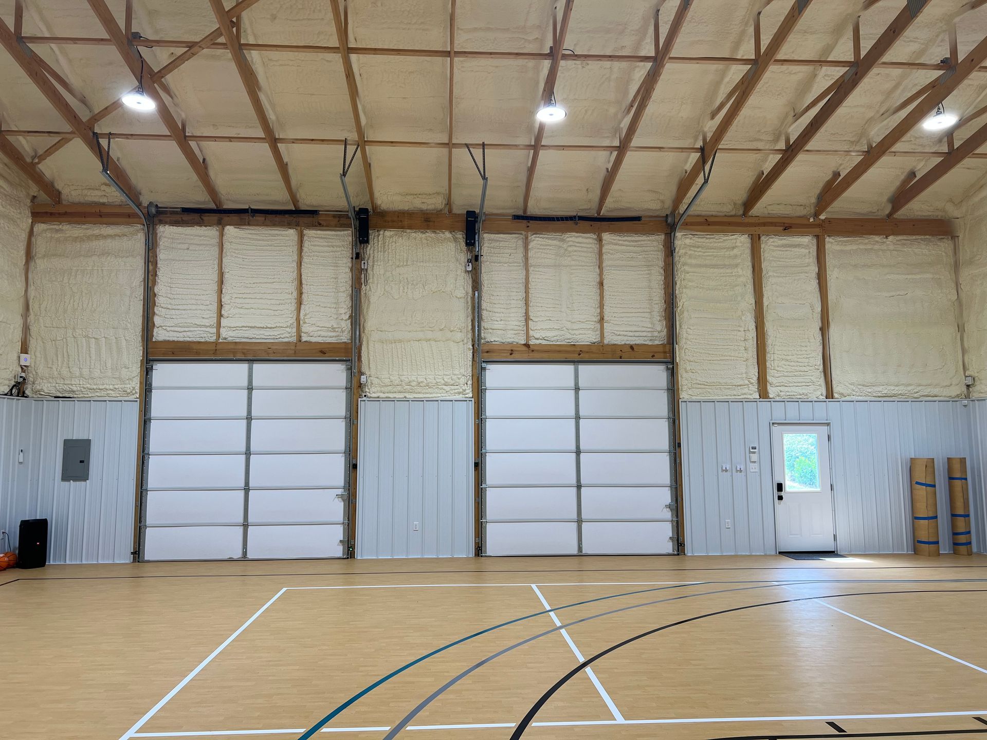 Indoor basketball court with two large white garage doors, a single entrance door, and foam-insulated walls and ceiling.