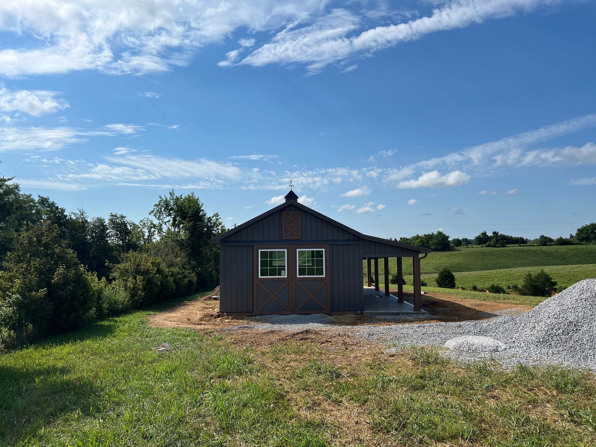 A dark-sided shed with two windows and a covered porch stands in a grassy field under a bright blue sky.