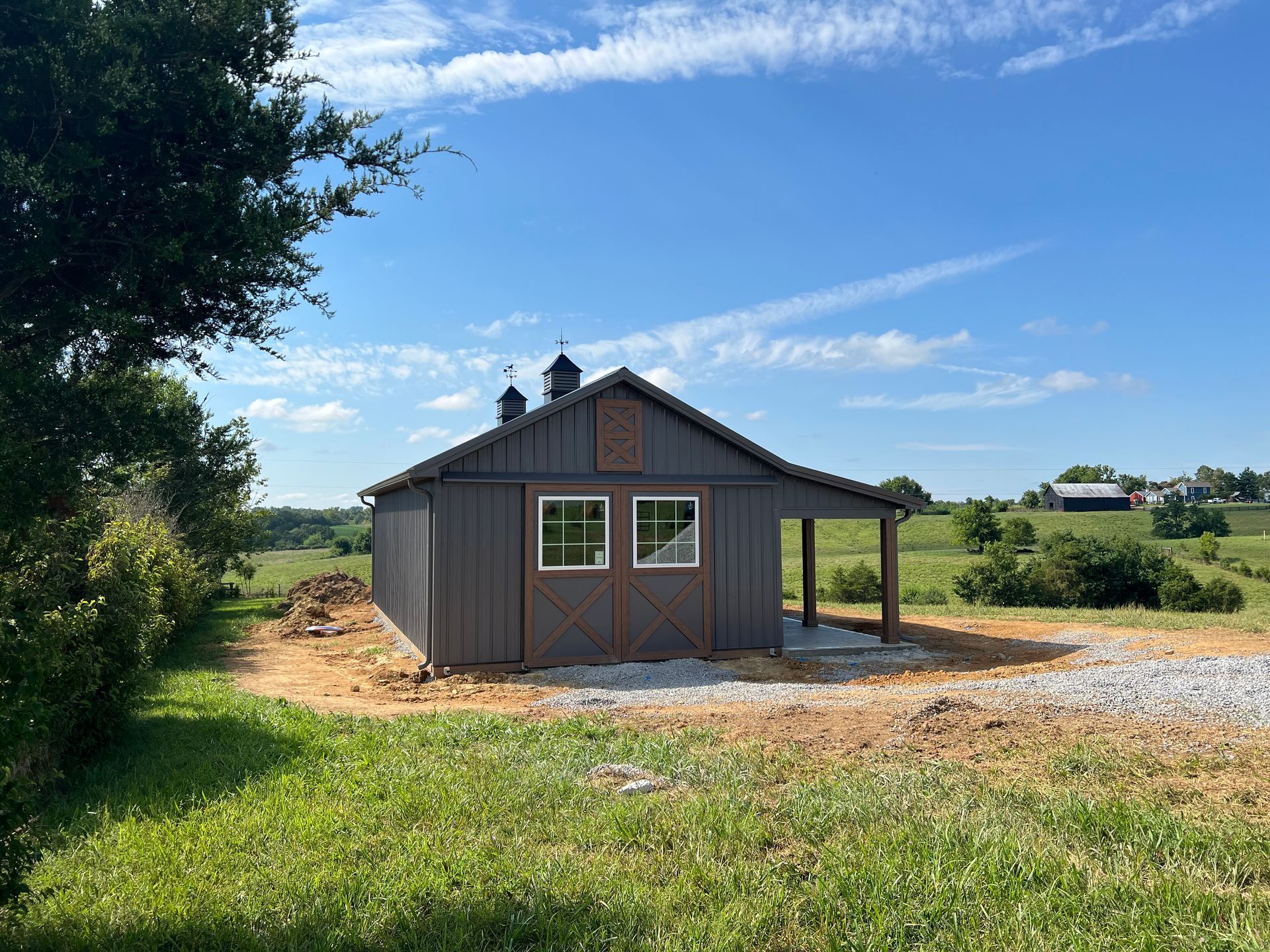 A dark gray, wood-sided building with two windows and a side porch, set in a sunny, grassy, rural landscape.