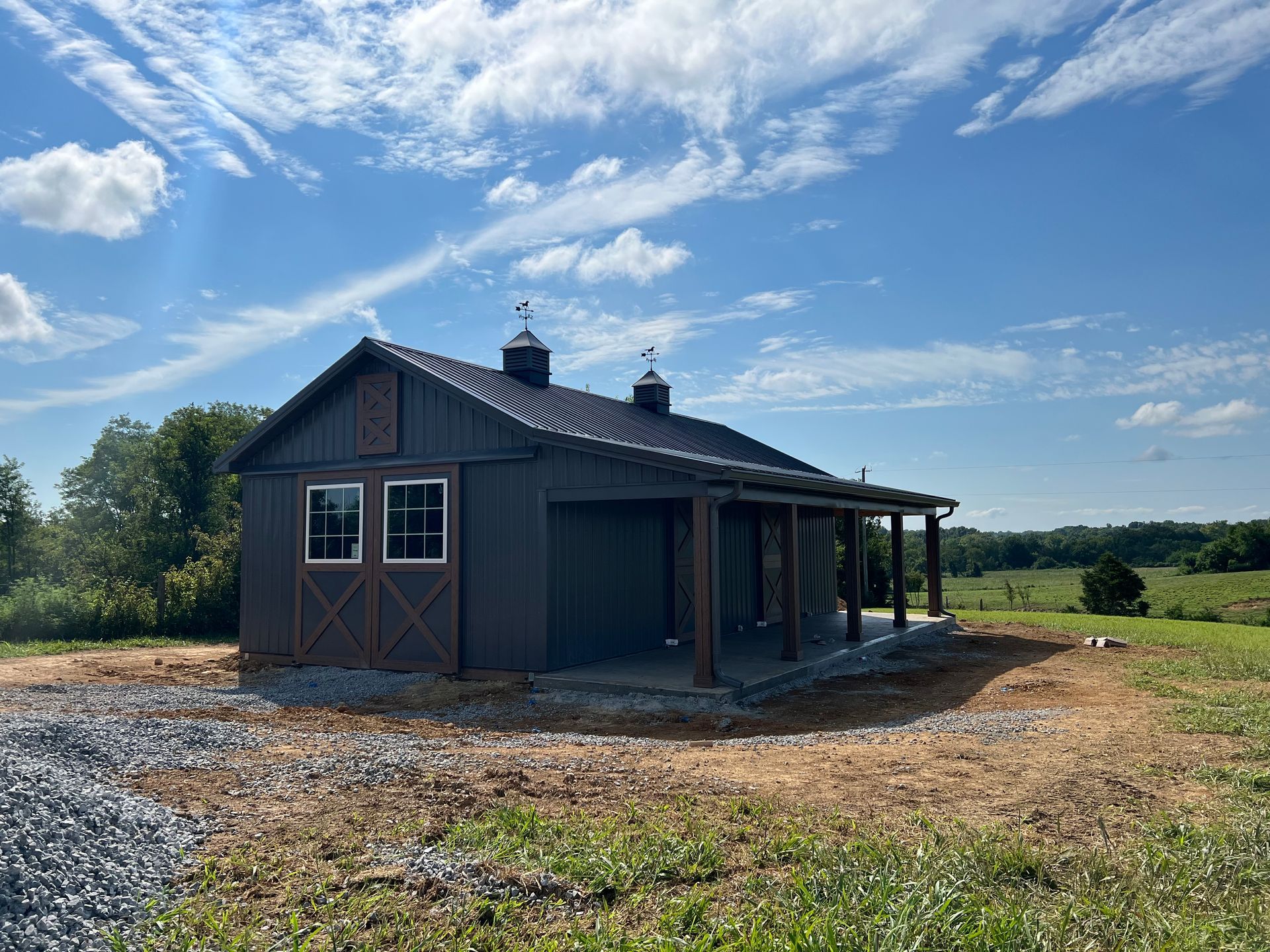 A dark gray barn with a porch and two cupolas, situated on a grassy landscape under a blue sky with scattered clouds.