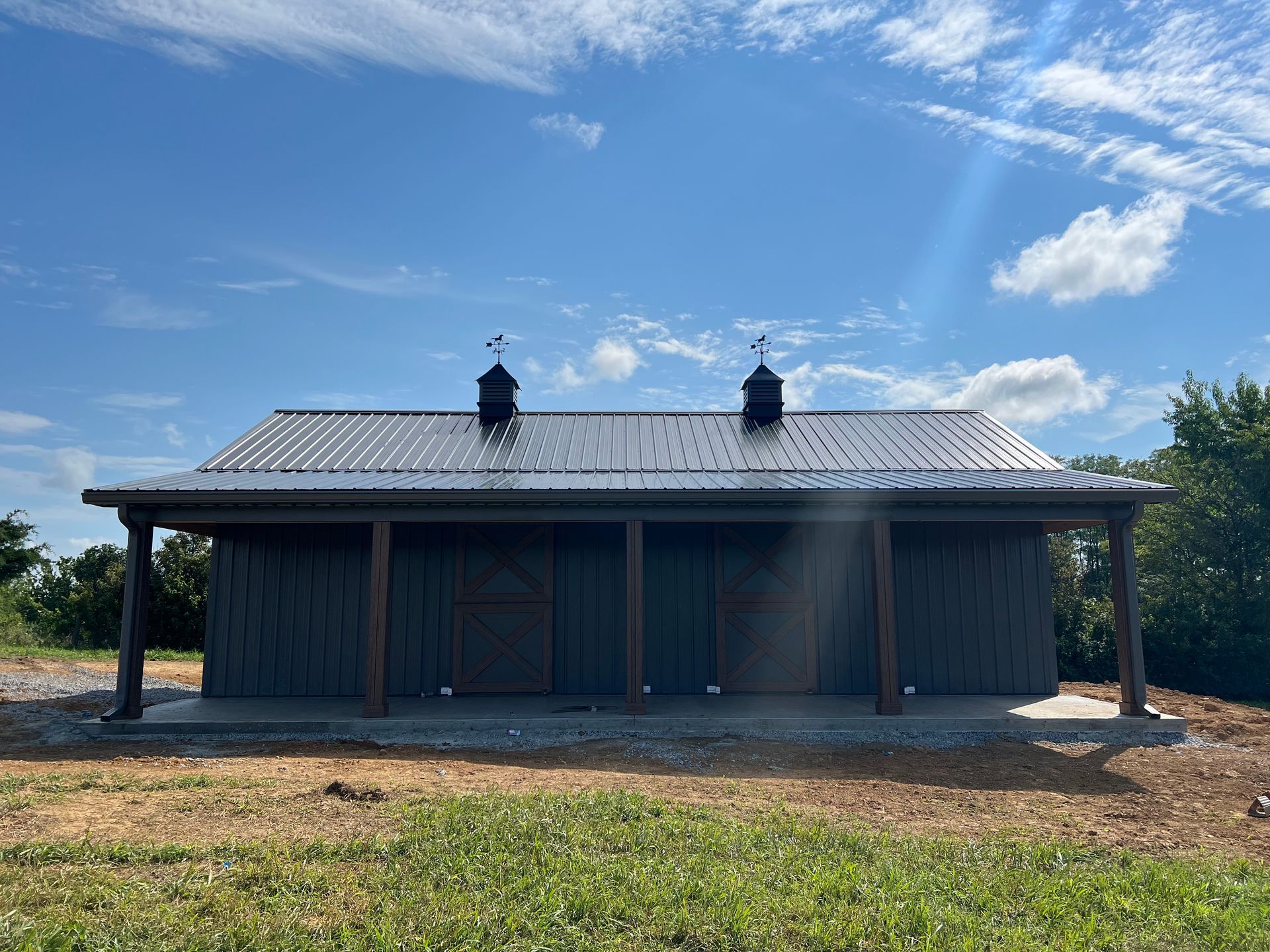 A dark, modern barn with a metal roof and two cupolas, situated on a patch of dirt and grass under a sunny blue sky.