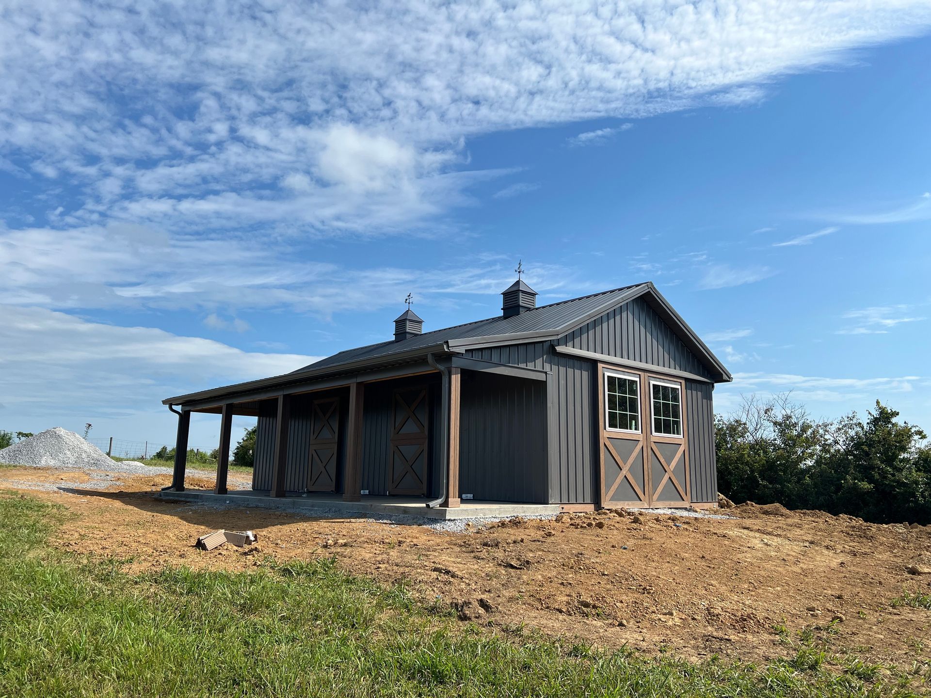 A dark gray barn with a porch and double sliding doors sits on a dirt lot under a bright blue, partially cloudy sky.