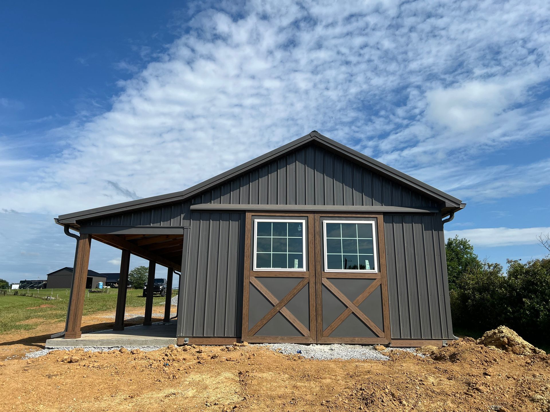 A dark gray shed with brown trim and a side porch, set against a bright, cloudy blue sky in a rural landscape.