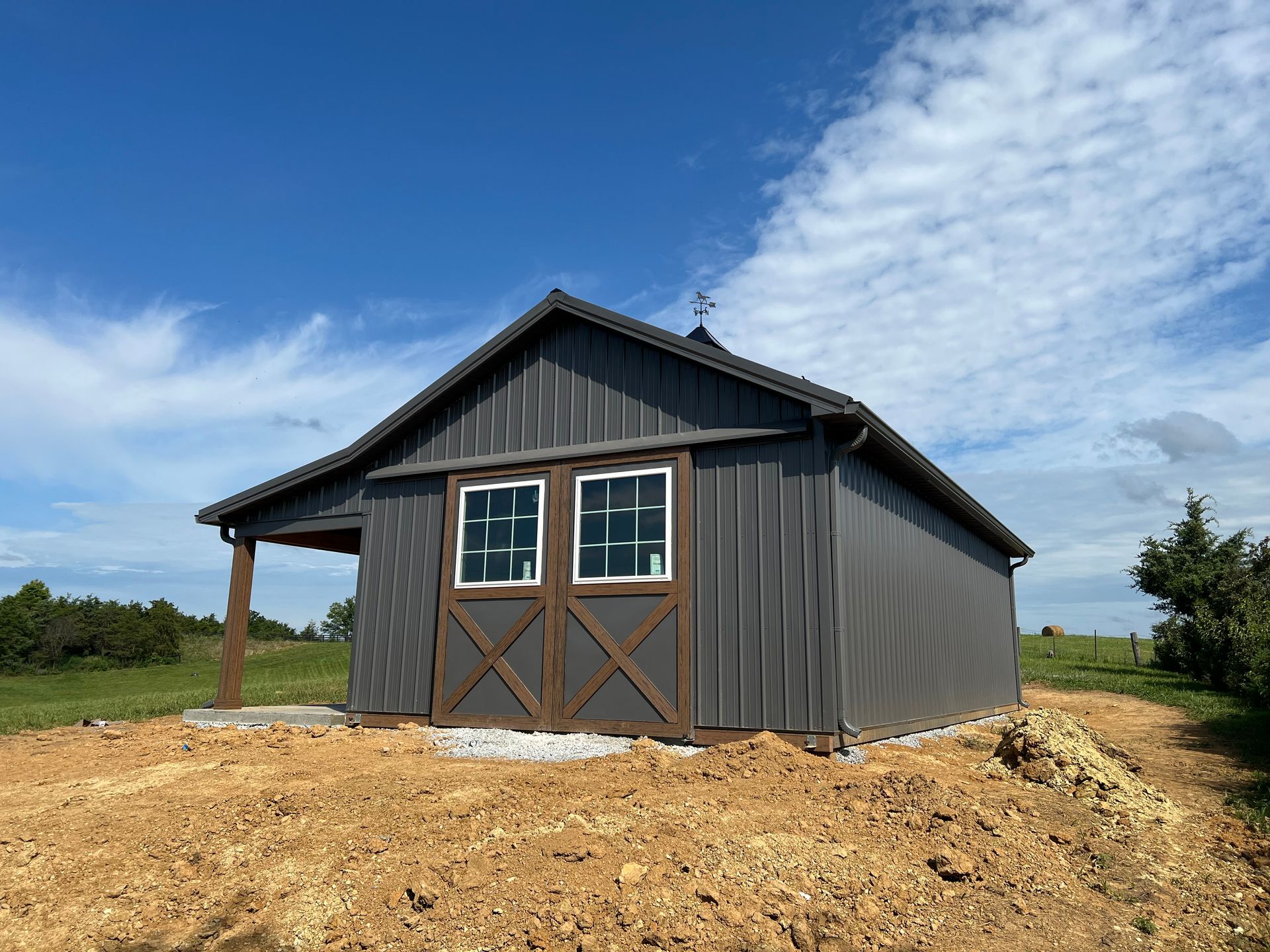 A gray metal barn with brown wooden trim and double windows sits on a dirt lot under a sunny blue sky.