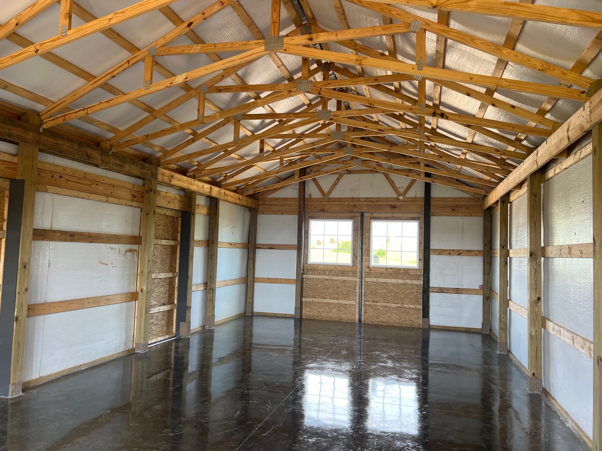 Interior view of a barn with wooden framing, exposed trusses, white insulation panels, and a polished concrete floor.