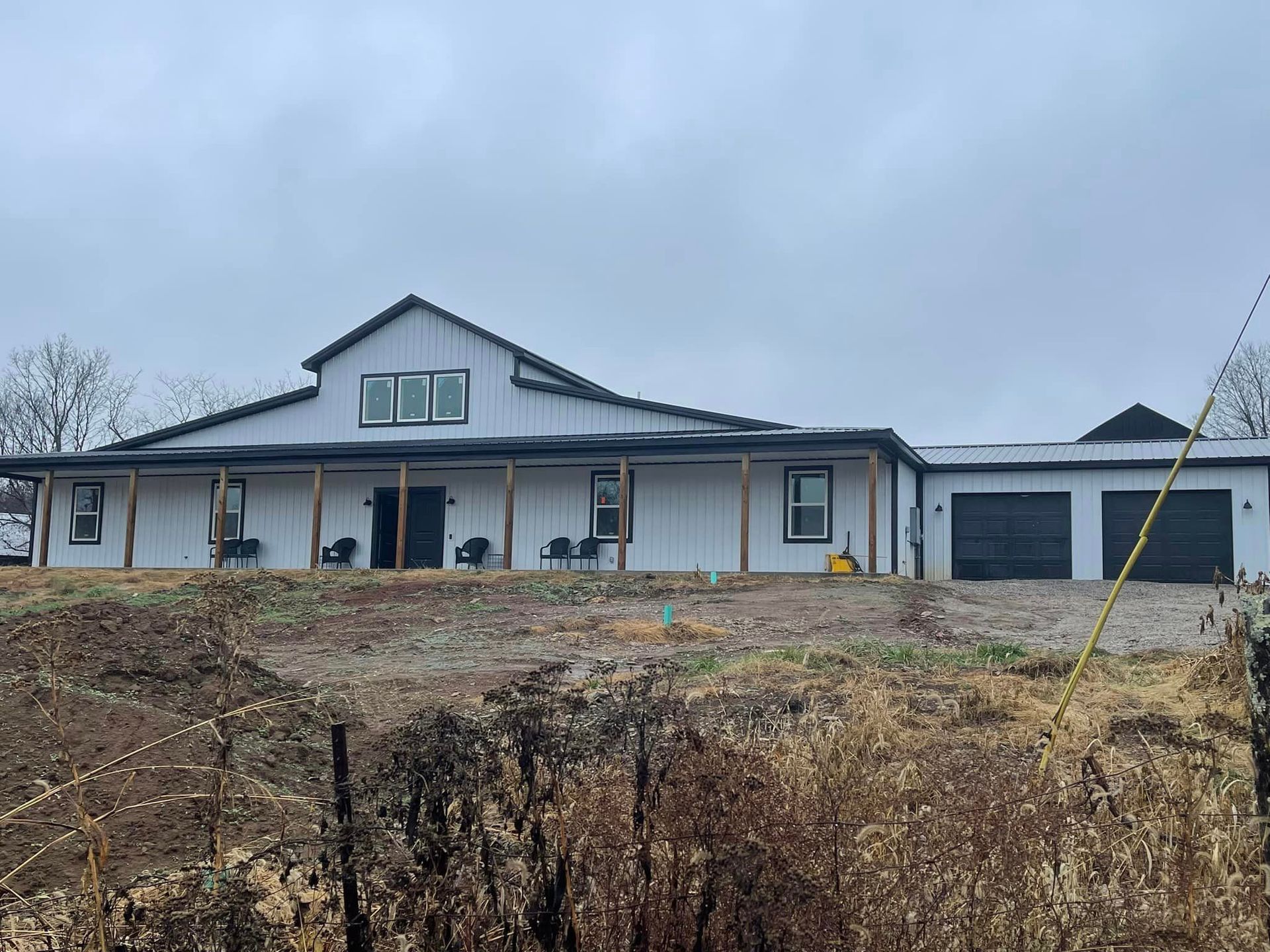 A wide, modern one-story white farmhouse with a metal roof, a wrap-around porch with wooden posts, and a two-car garage.
