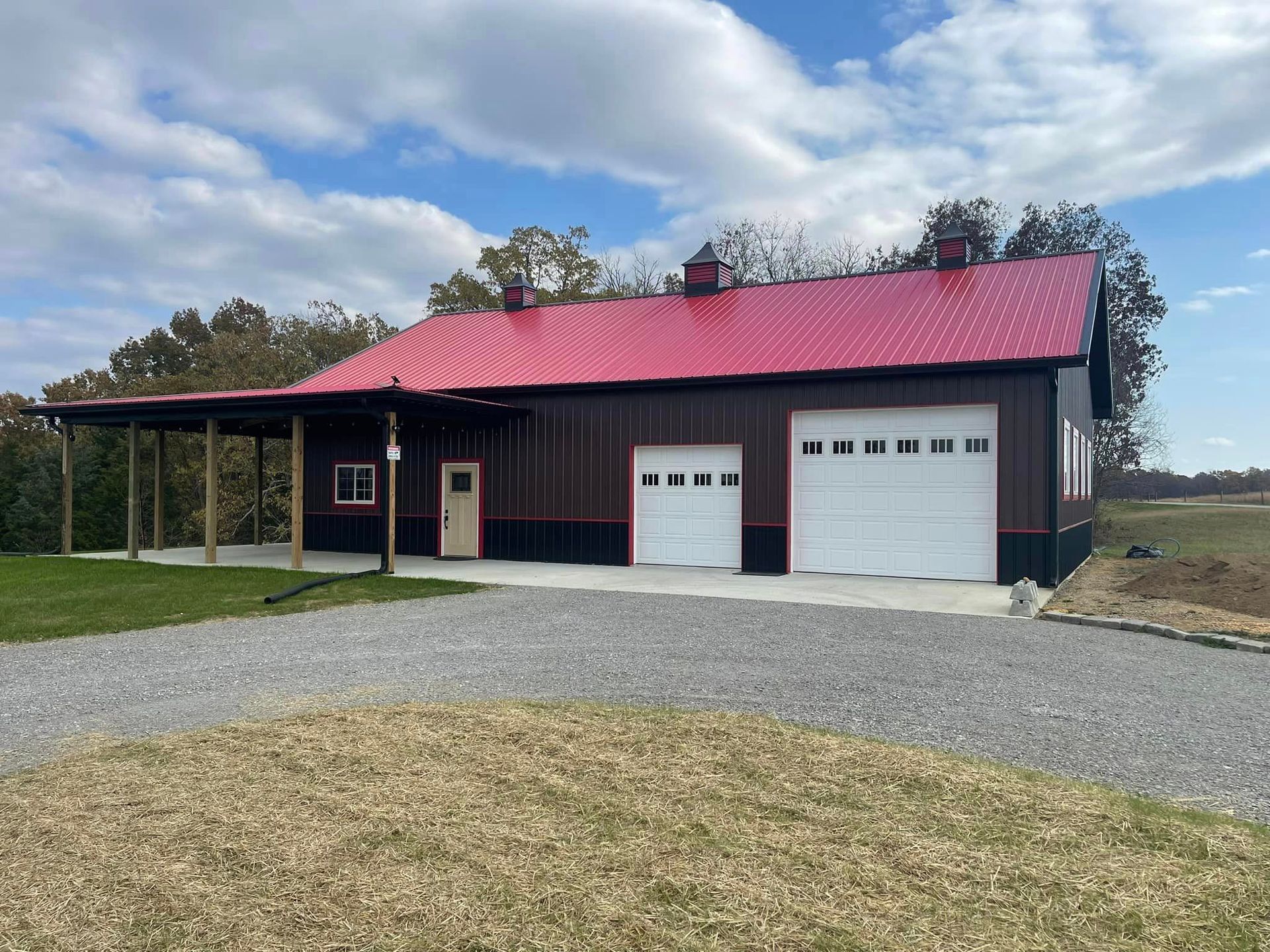 A modern brown metal building with a bright red roof, white garage doors, and a front porch, set on a gravel driveway.