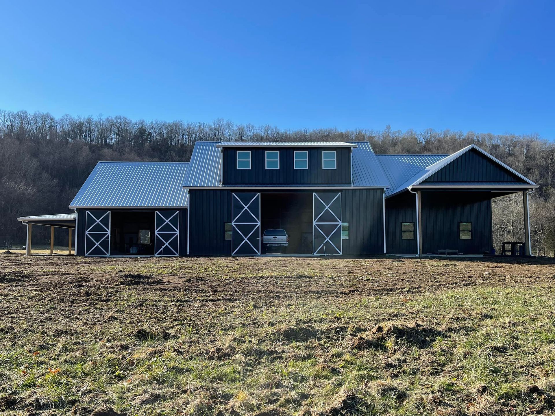 A dark-sided barn with a metal roof and white x-patterned doors sits in a muddy, rural field against a treeline.