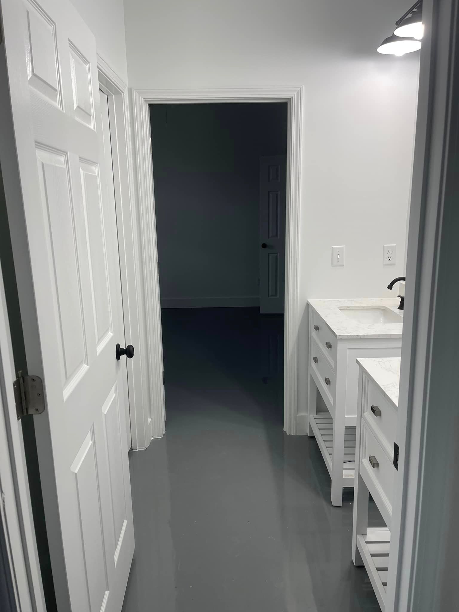 A view from a doorway into a bathroom with white cabinets, a gray floor, and an open door leading to another room.