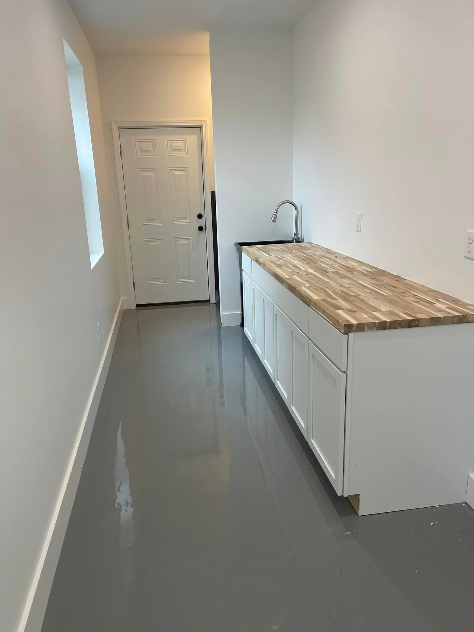 A narrow room with a grey, glossy floor, white walls, a single door at the end, and a counter with a sink.