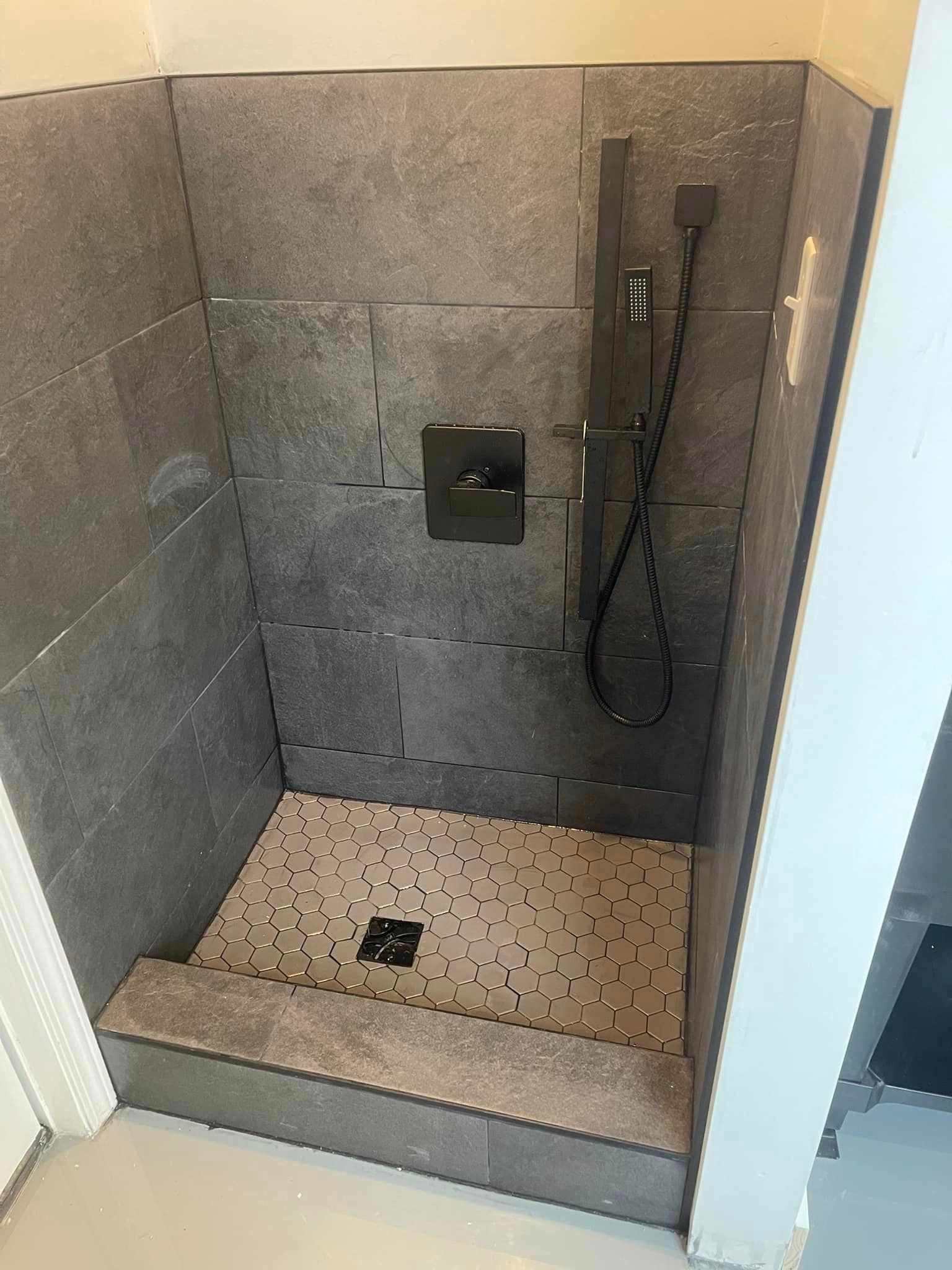 A walk-in shower with dark gray stone-look tiles, a black shower fixture, and a light-colored pebbled floor.