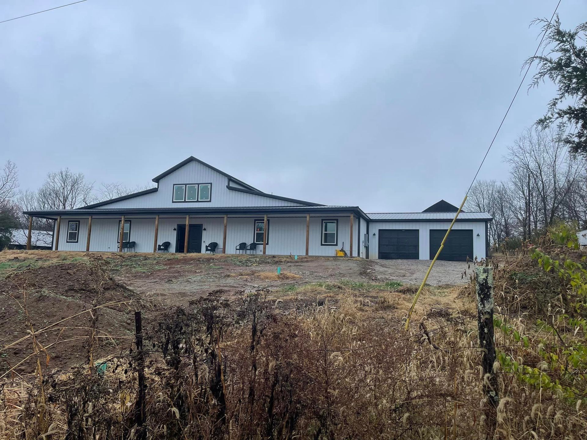 A wide, white, modern farmhouse with a dark roof and garage, set behind a rural fence on an overcast day.