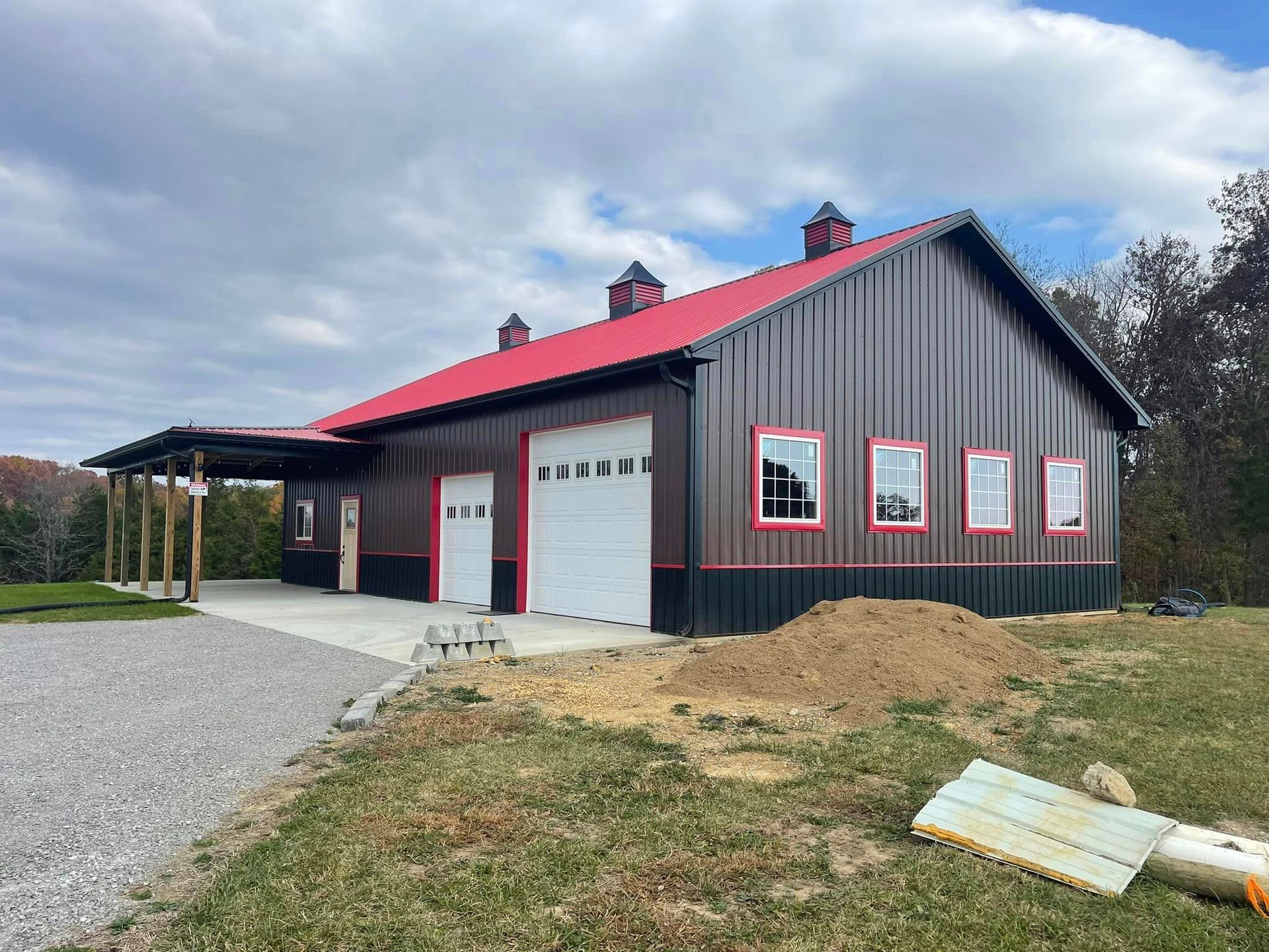 A modern, dark-sided barn with a red metal roof, white garage doors, and a covered side porch under a partly cloudy sky.