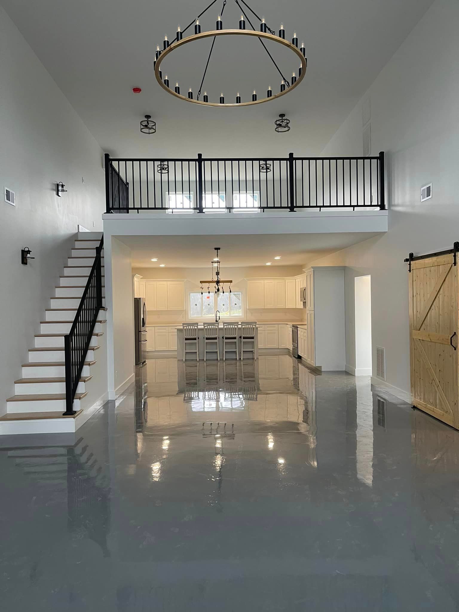 Modern, high-ceilinged interior with a loft, gray epoxy flooring, a black-railed staircase, and a large chandelier.