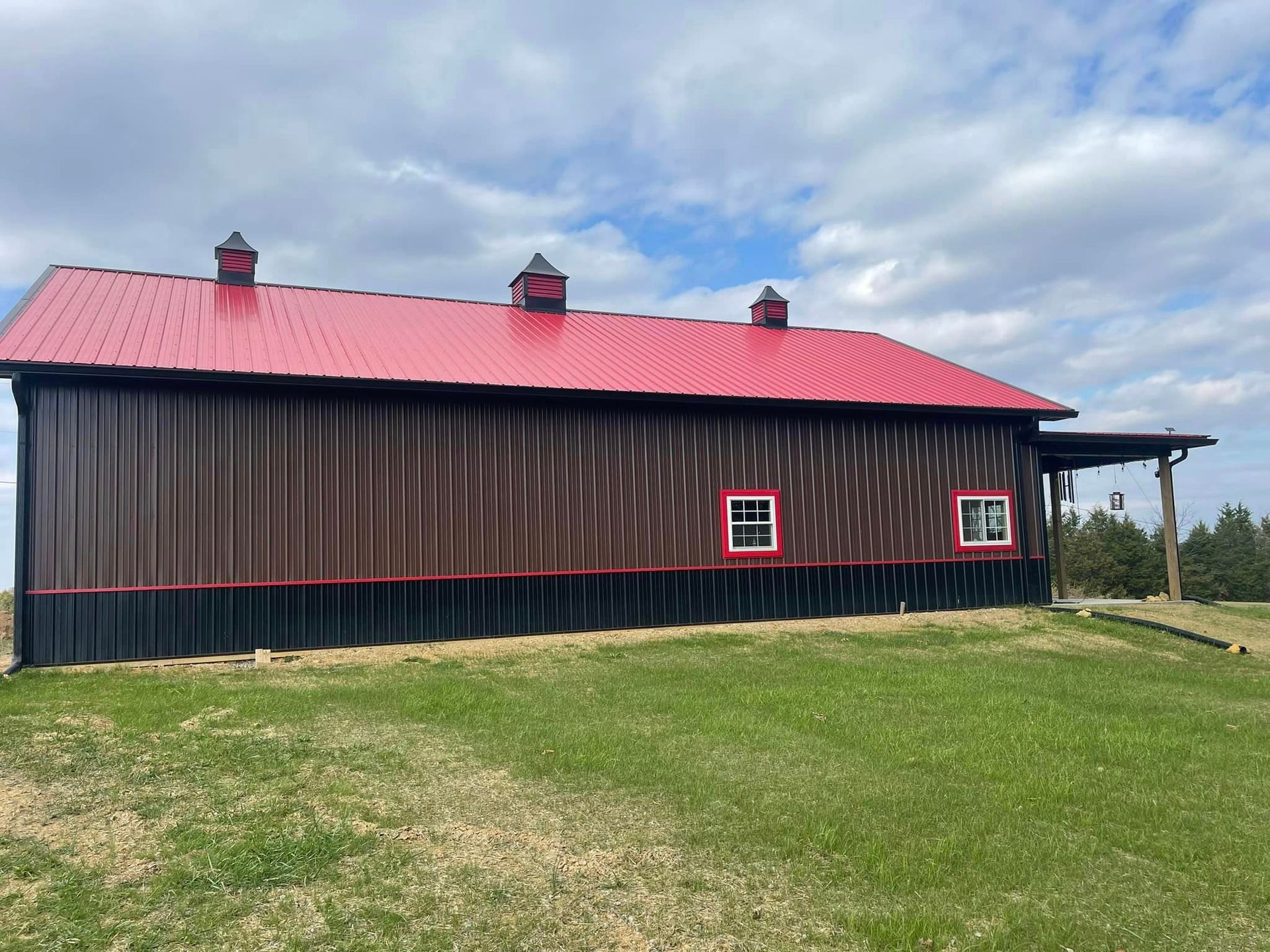 A long barn with a red metal roof, brown corrugated siding, a black lower trim, and three cupolas under a cloudy sky.