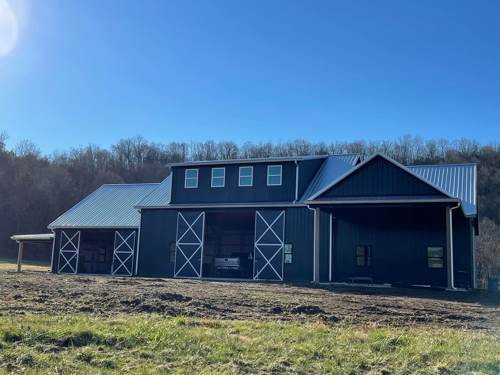 A large, dark blue barn with white cross-braced sliding doors stands on a dirt lot under a bright blue sky.