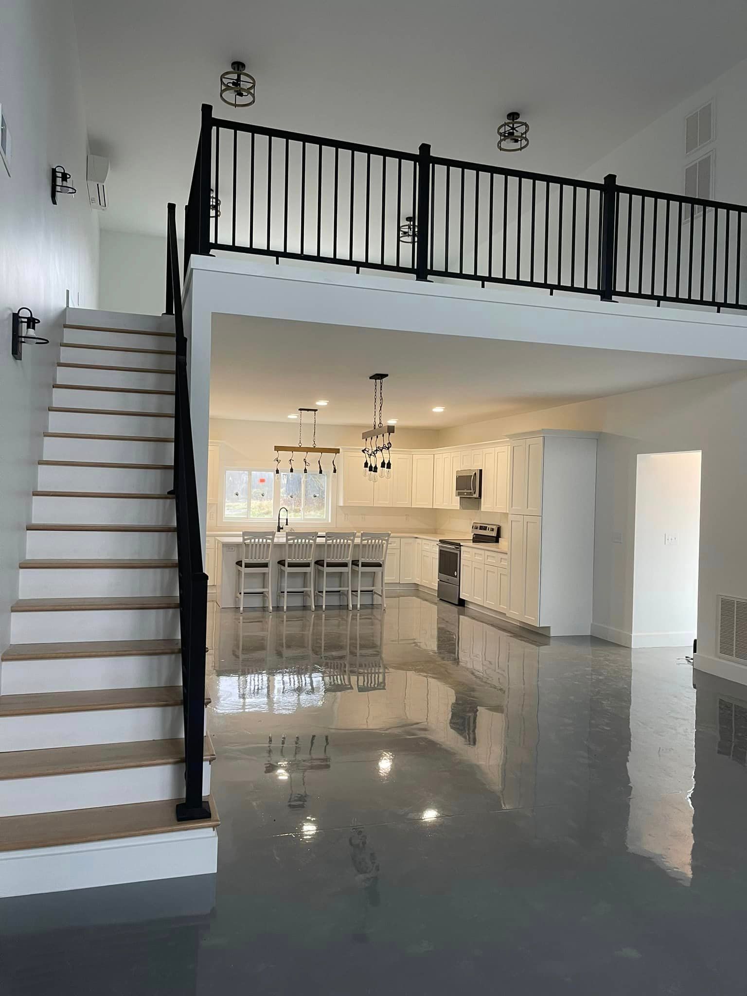 An open-concept interior with a glossy gray floor, a staircase with black railings, and a loft overlooking a kitchen.