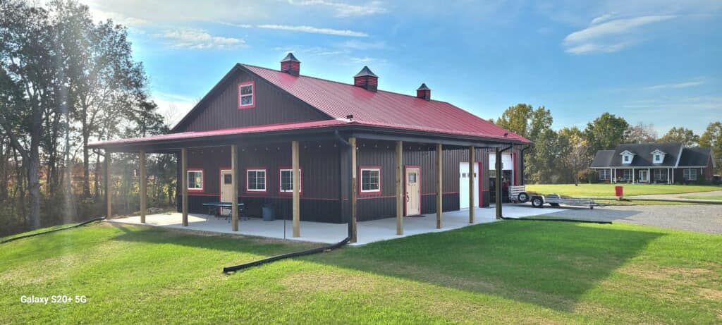 A rustic, dark-brown barn with a bright red metal roof, white trim, and a wrap-around porch set on a grassy lawn.