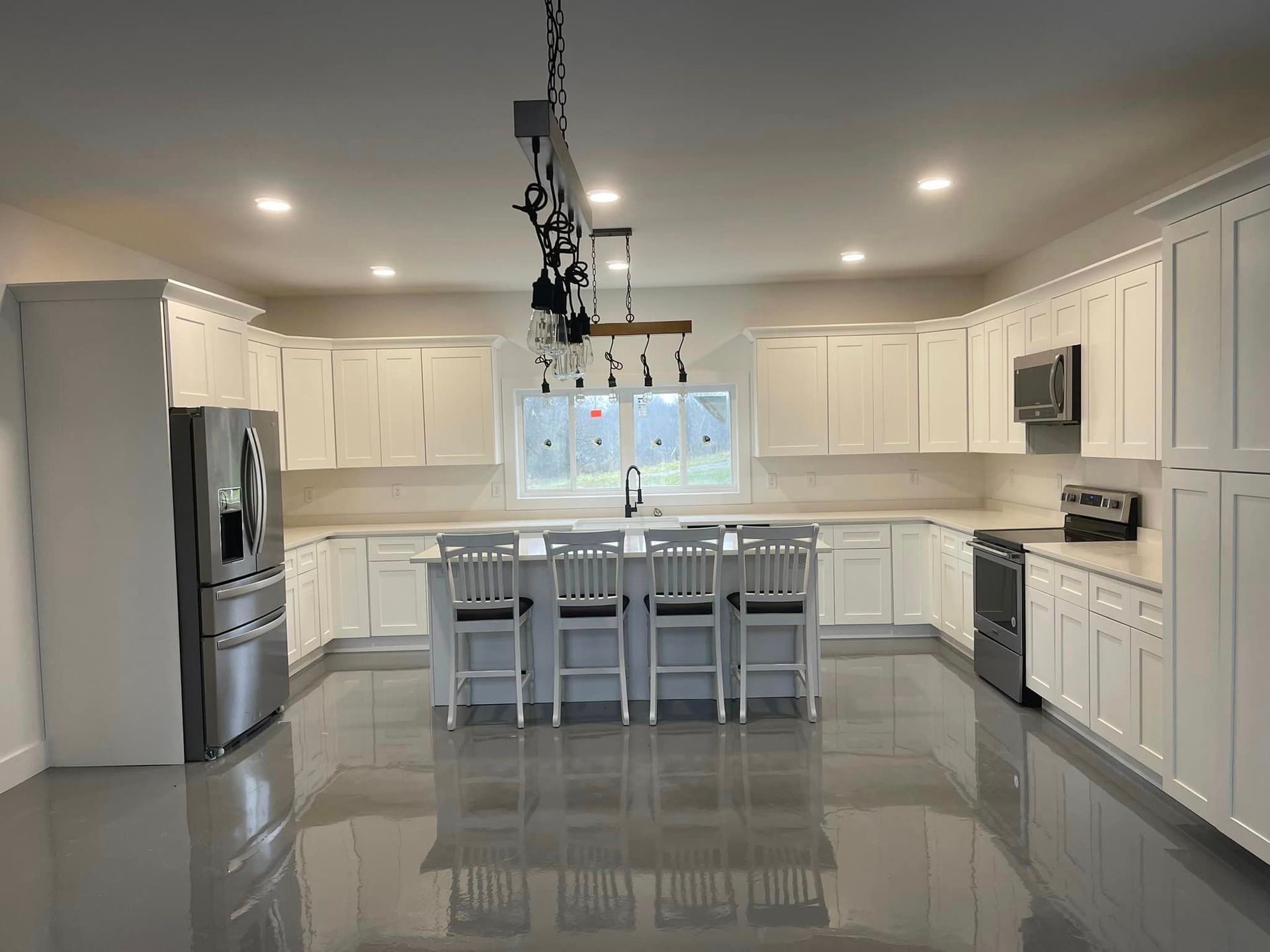 A spacious kitchen with white cabinets, stainless steel appliances, a central island with four chairs, and grey flooring.