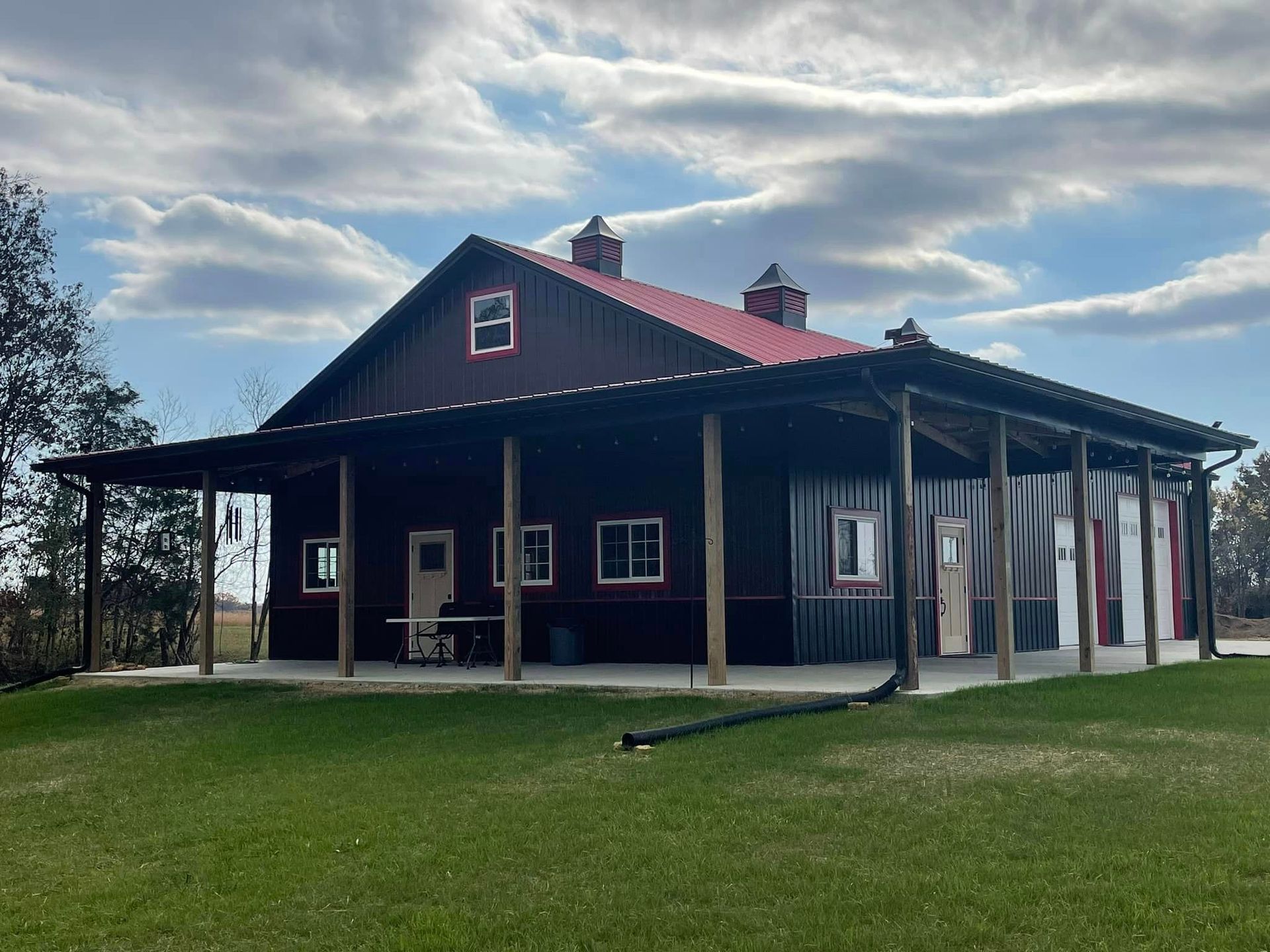 A dark brown barn with a red metal roof and a wide wrap-around porch, set against a cloudy blue sky and green grass.