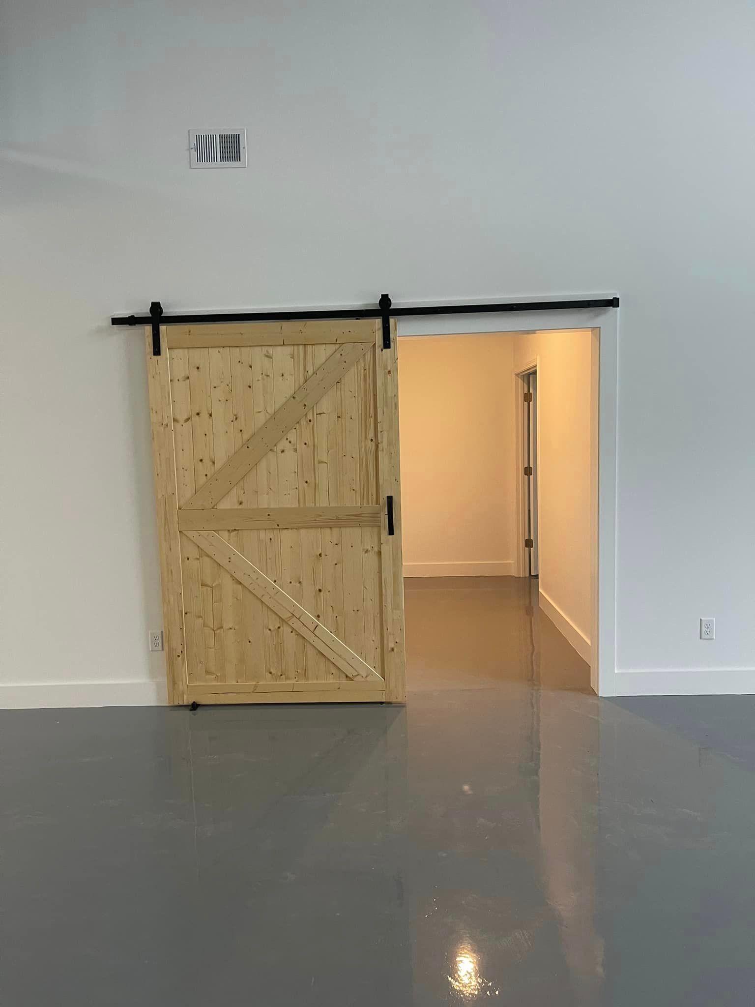 A natural-wood sliding barn door on a black metal track, partially open to reveal a room with grey flooring and white walls.