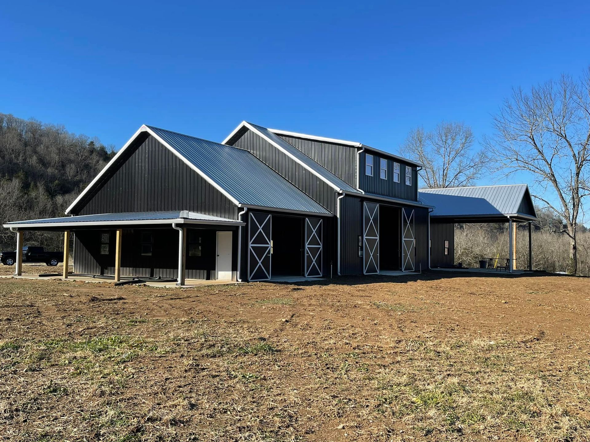 A modern, dark gray pole barn with a metal roof and large sliding barn doors sits on a dirt lot under a clear blue sky.