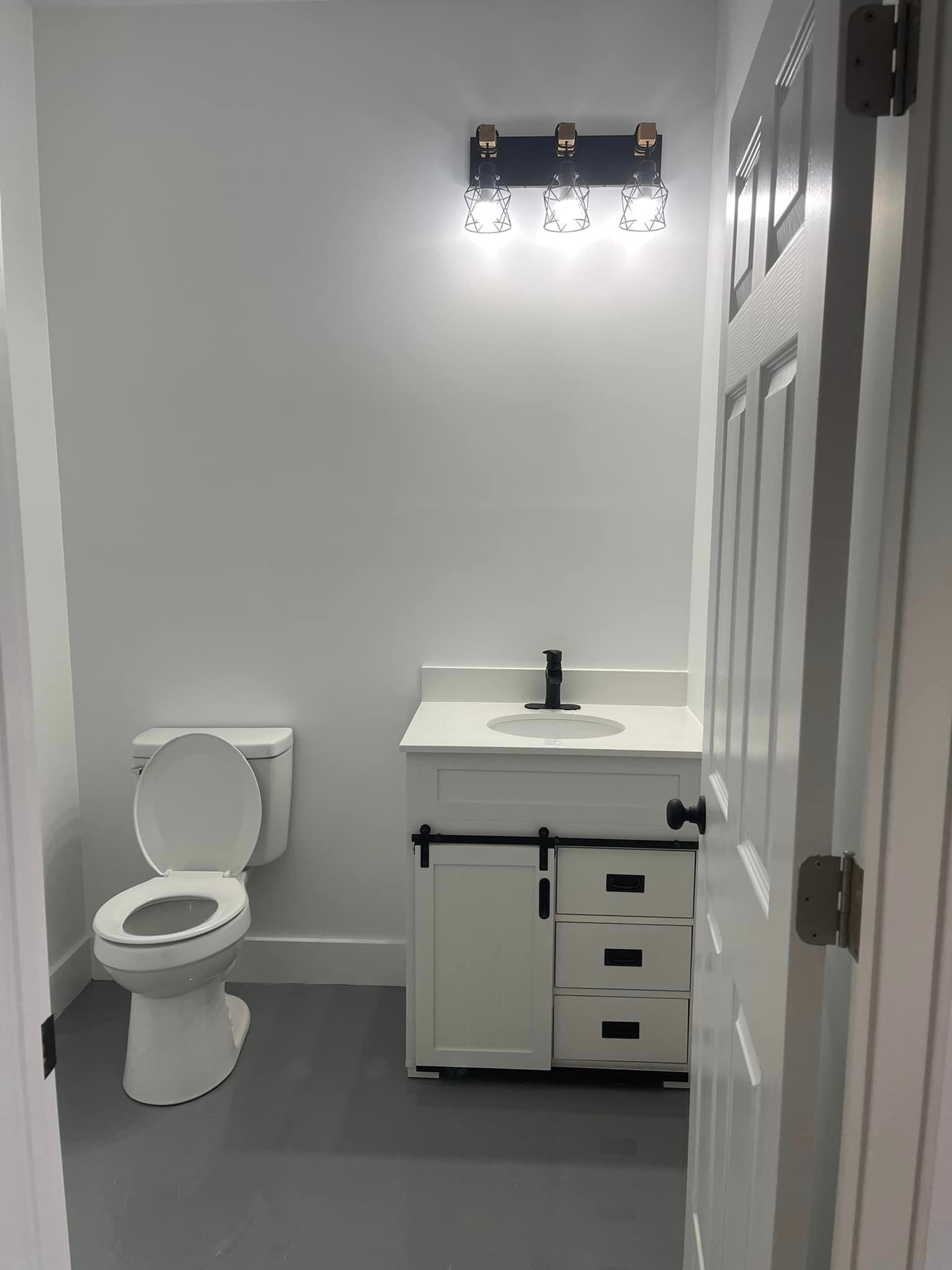 A small bathroom with a white toilet and a white vanity with black hardware and a dark light fixture.