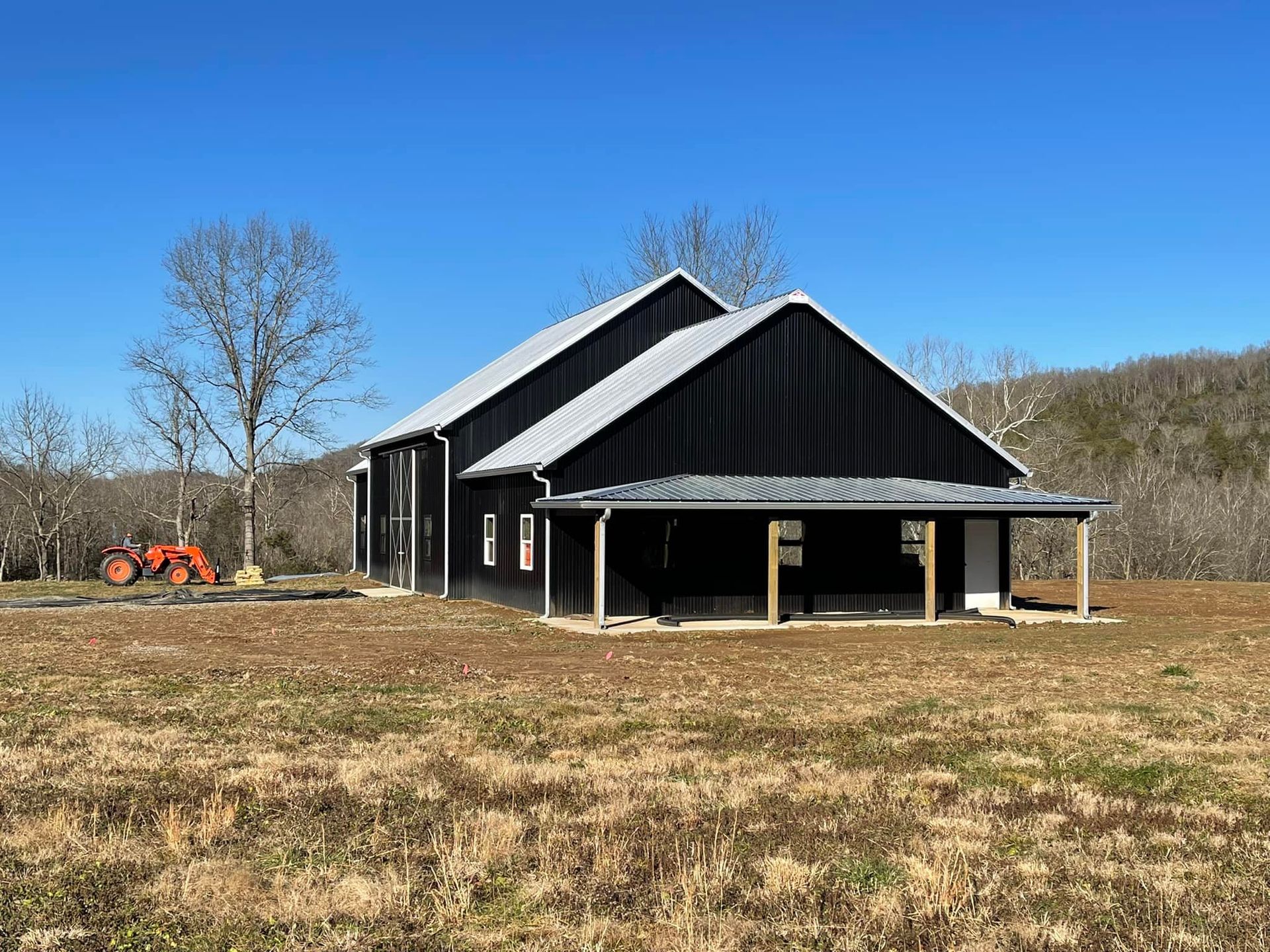 A black barn with a silver metal roof and wrap-around porch stands in a dry, open field under a clear blue sky.