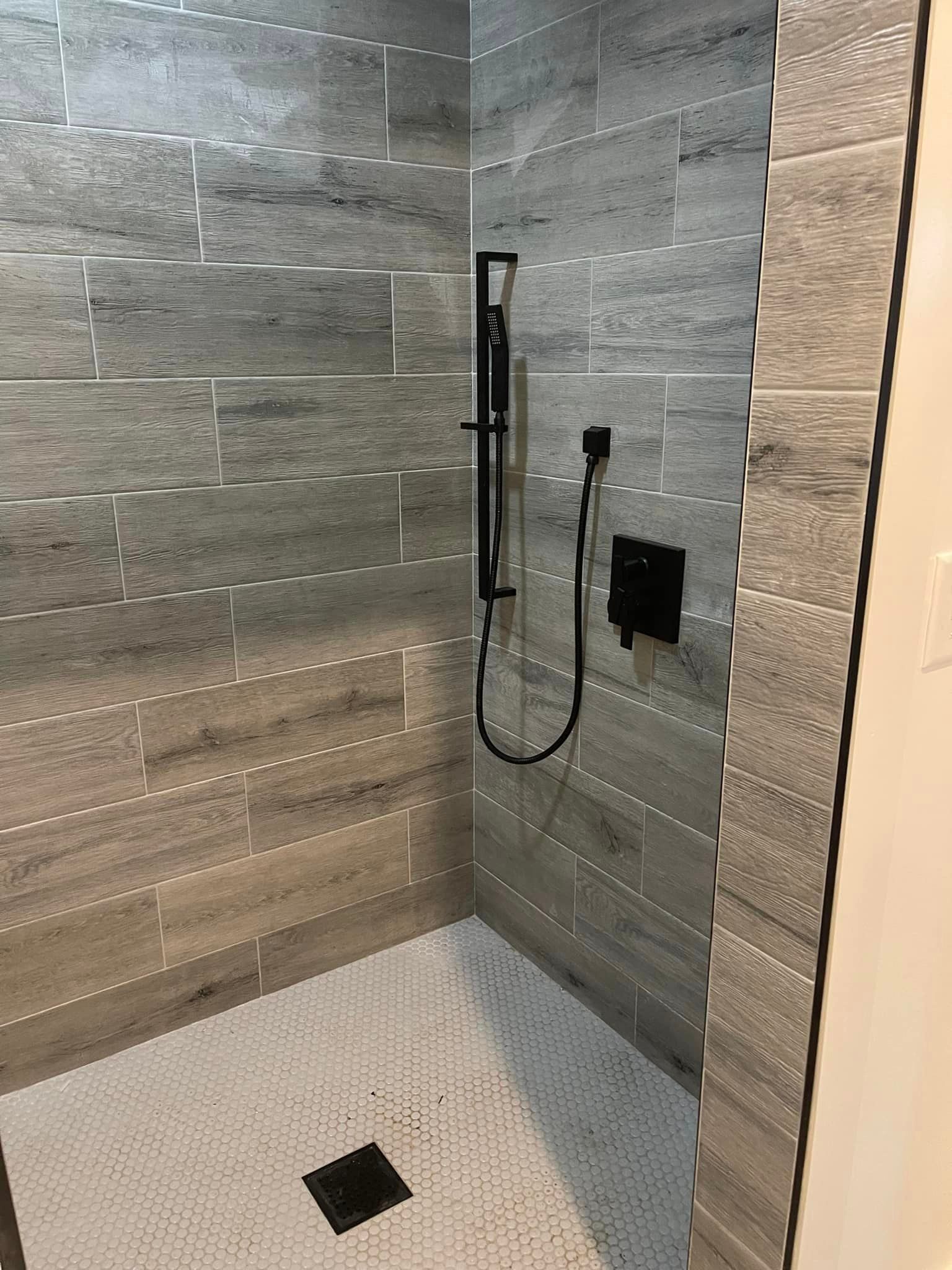 A walk-in shower with horizontal gray wood-look wall tiles, a small white hexagonal tile floor, and matte black fixtures.