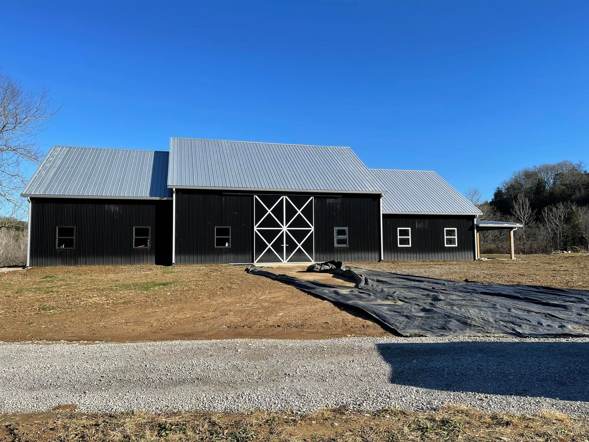 A black barn with a metal roof and large white cross-buck double doors, set in a field under a clear blue sky.