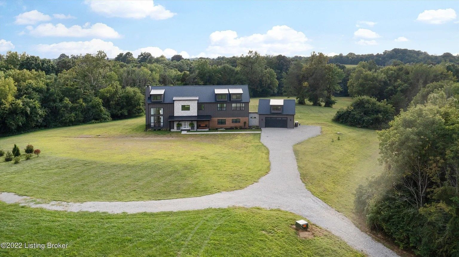 An aerial view of a multi-story home with a gravel driveway, surrounded by grassy fields and mature trees under a blue sky.