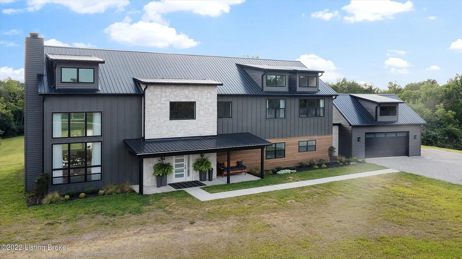A modern, two-story house with dark gray siding, a stone facade center, metal roof, and attached garage in a grassy yard.