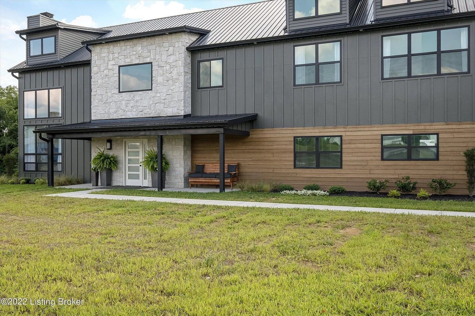A modern two-story home with a stone and dark vertical metal exterior, wood accents, a front porch, and a grassy lawn.