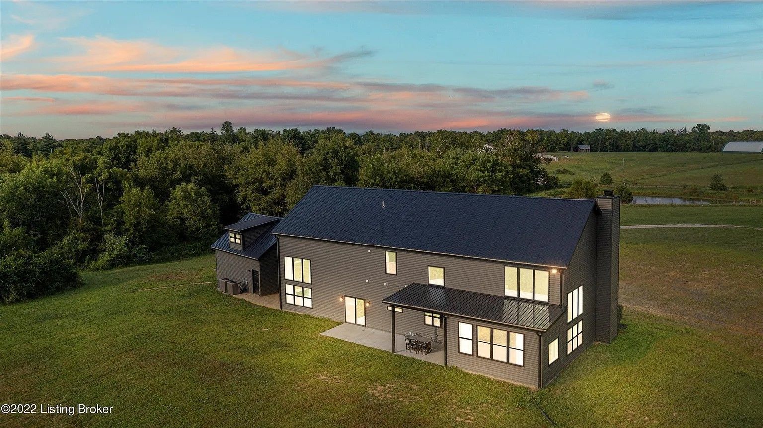 An aerial view of a dark-sided, modern two-story home in a grassy field during a vibrant, colorful sunset.