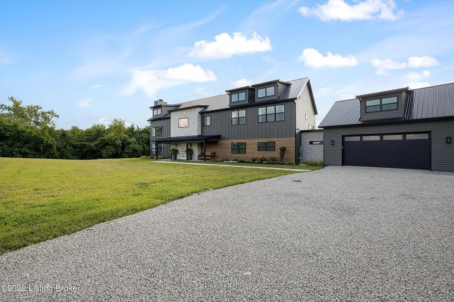 A large, modern home with a grey metal exterior, dormer windows, and a detached two-car garage sits beside a gravel drive.