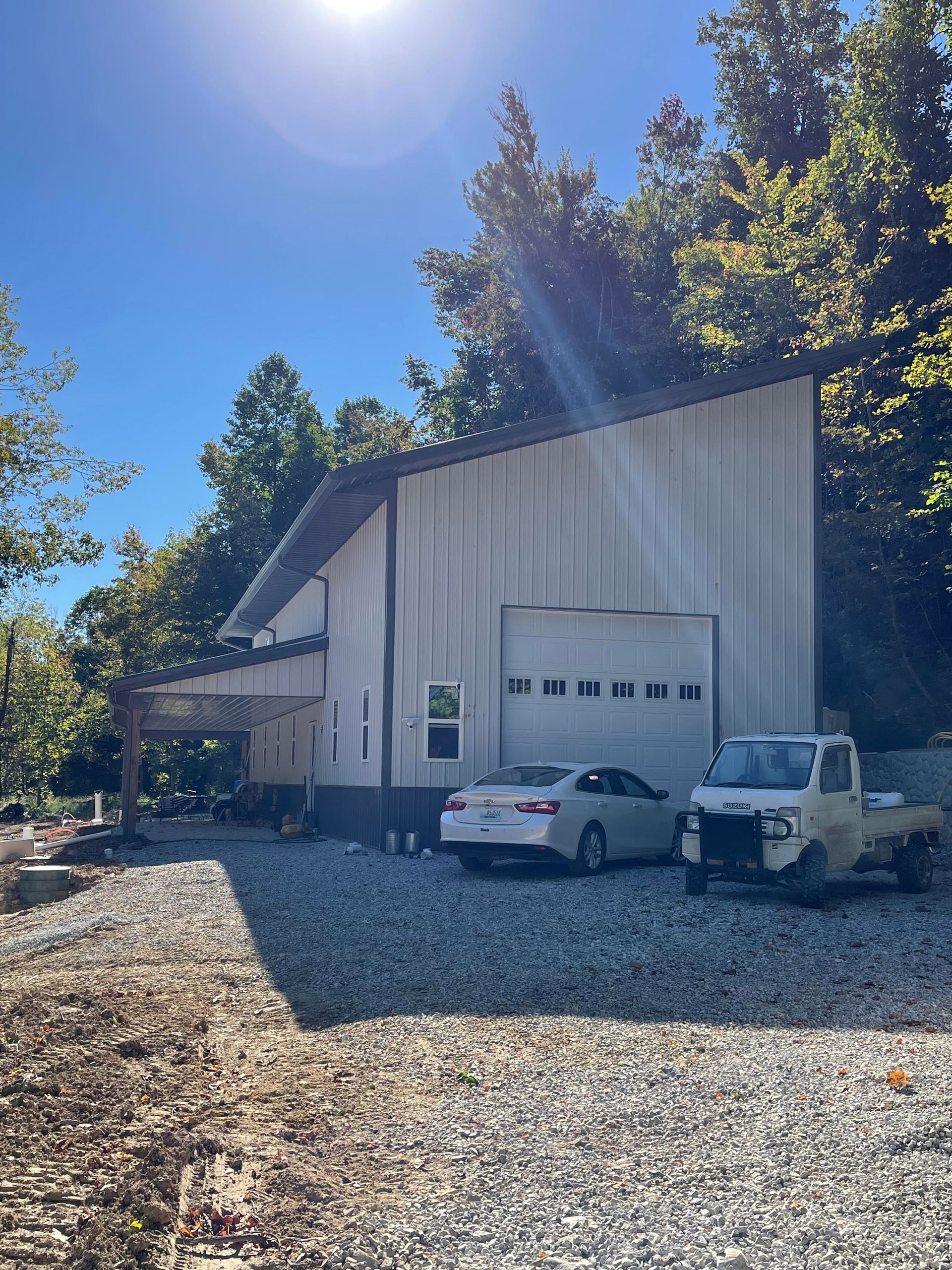 A white metal workshop with a garage door and side porch, located on a gravel lot under a bright blue sky.