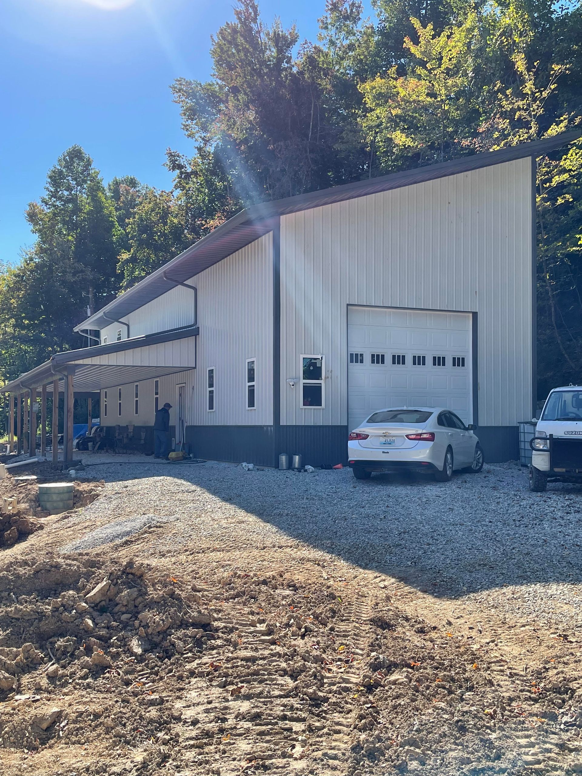 A white metal-sided building with a shed roof and a garage door, parked next to a white car on a gravel lot.