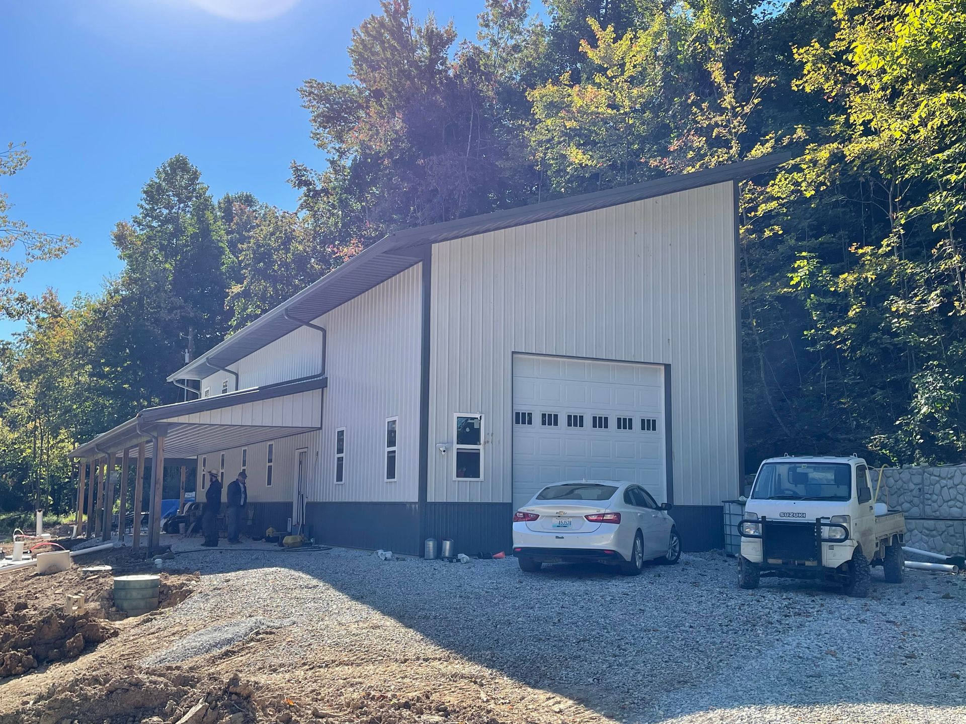 A modern metal building with a shed roof, a white roll-up door, a covered porch, and vehicles parked on a gravel lot.