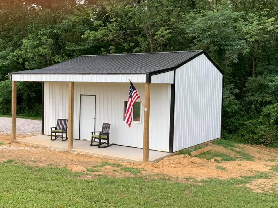 A white metal shed with a black roof and porch, featuring two rocking chairs, a door, a window, and an American flag.