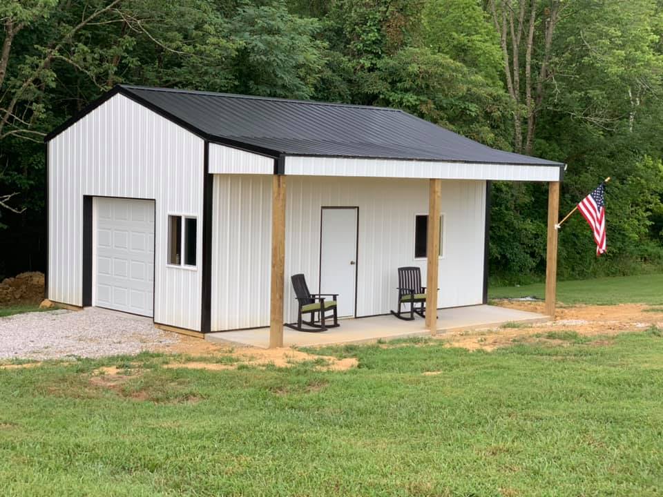 A white metal workshop with a black roof and a covered porch, featuring a garage door, a side entry door, and rocking chairs.
