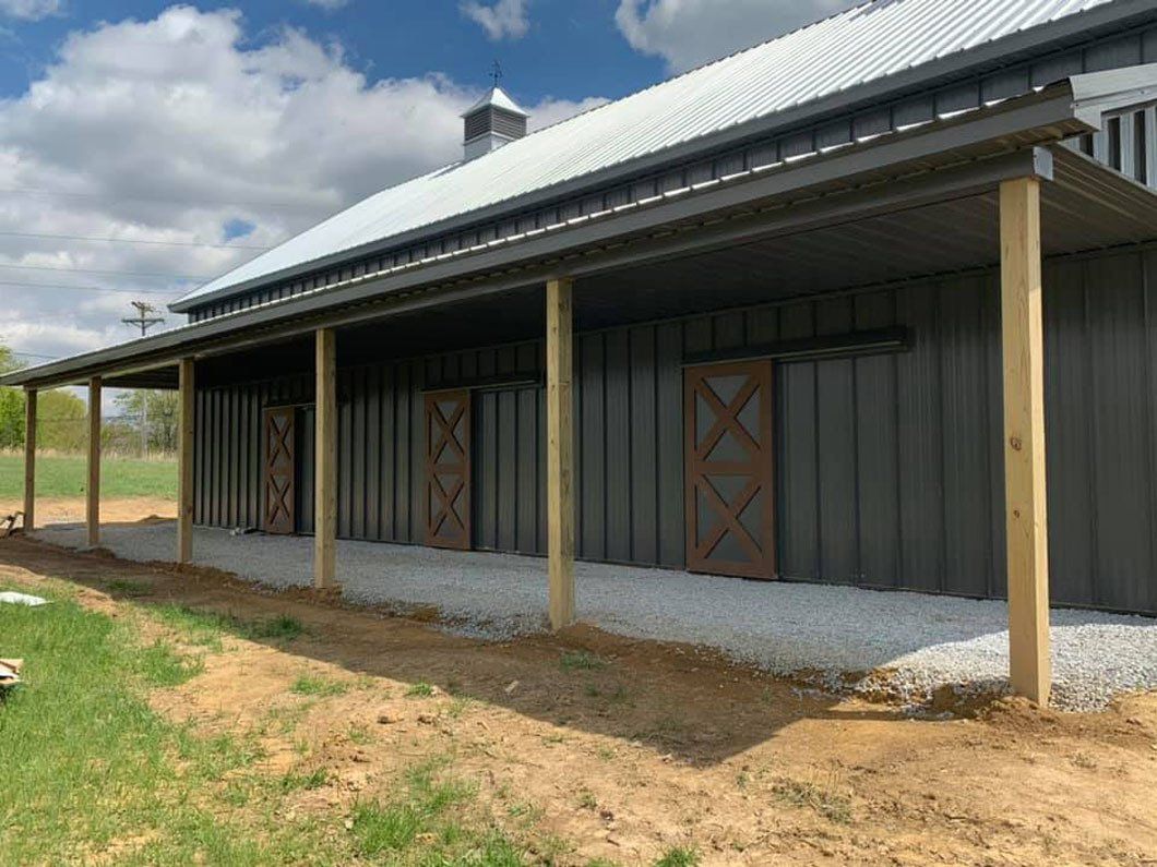 A modern dark metal barn with a long covered porch, wooden support posts, and wooden sliding doors over a gravel path.