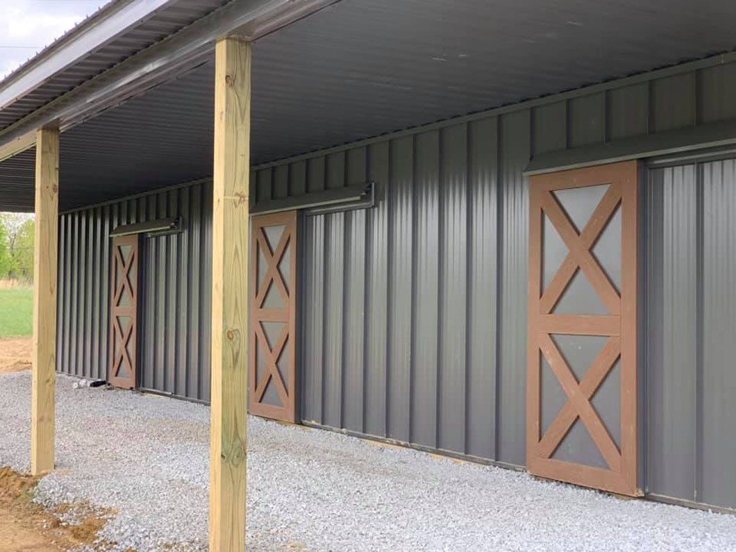 Gray metal wall of a building with three rustic, brown wooden barn doors and a gravel path under a porch overhang.