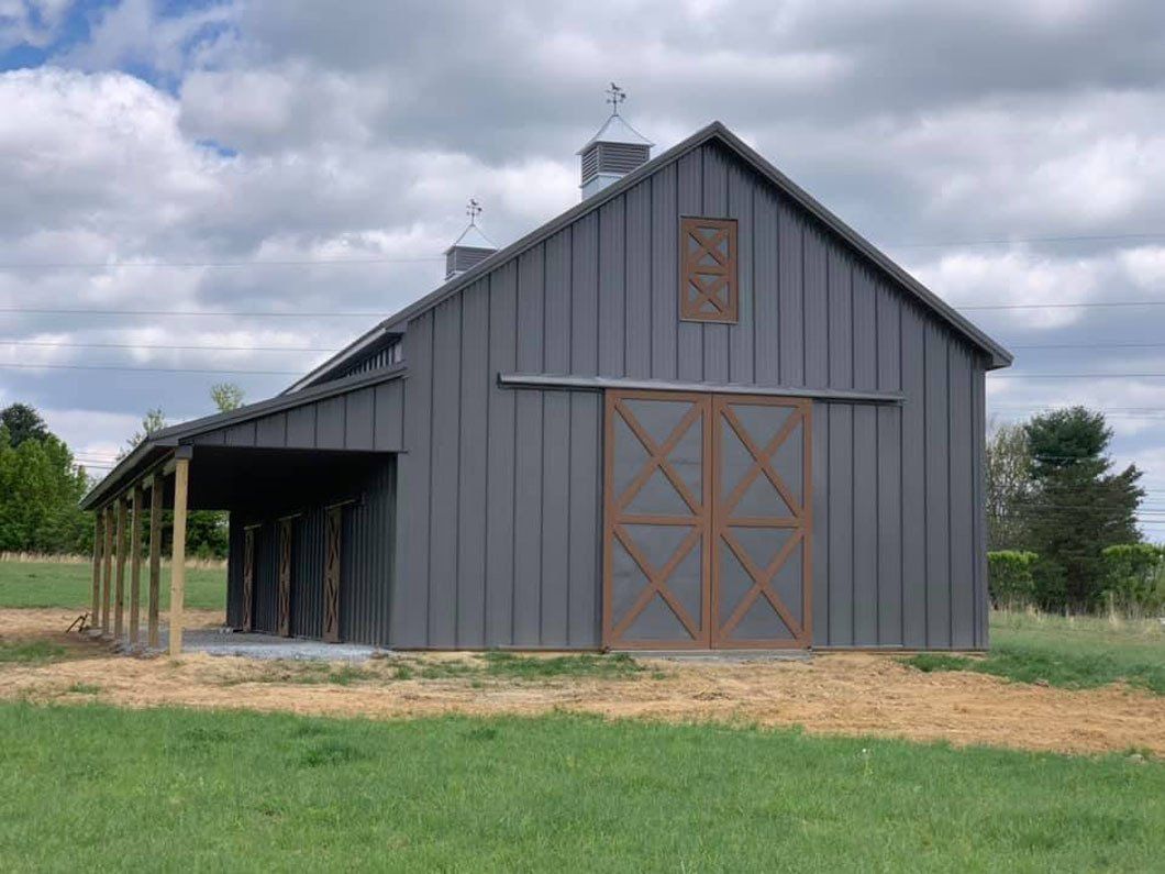 A grey barn with a side lean-to and brown cross-buck sliding doors, set in a grassy field under a cloudy sky.