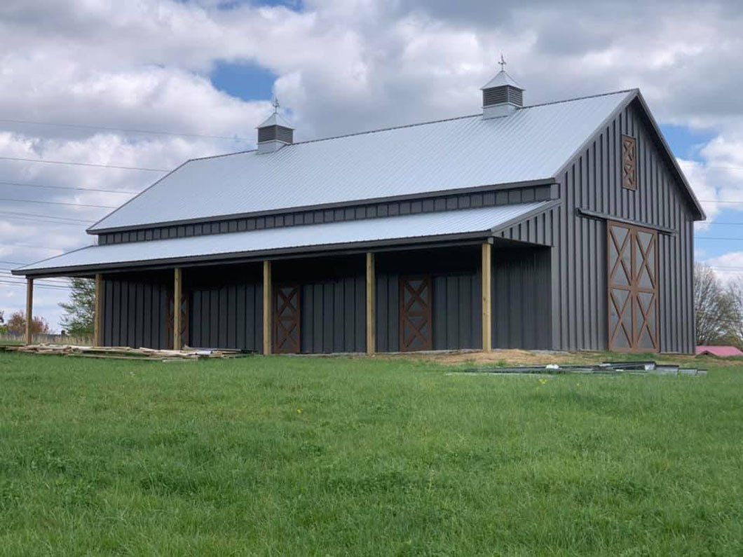 A modern dark gray metal barn with a covered porch, wooden doors, and two cupolas, set in a green field under a blue sky.