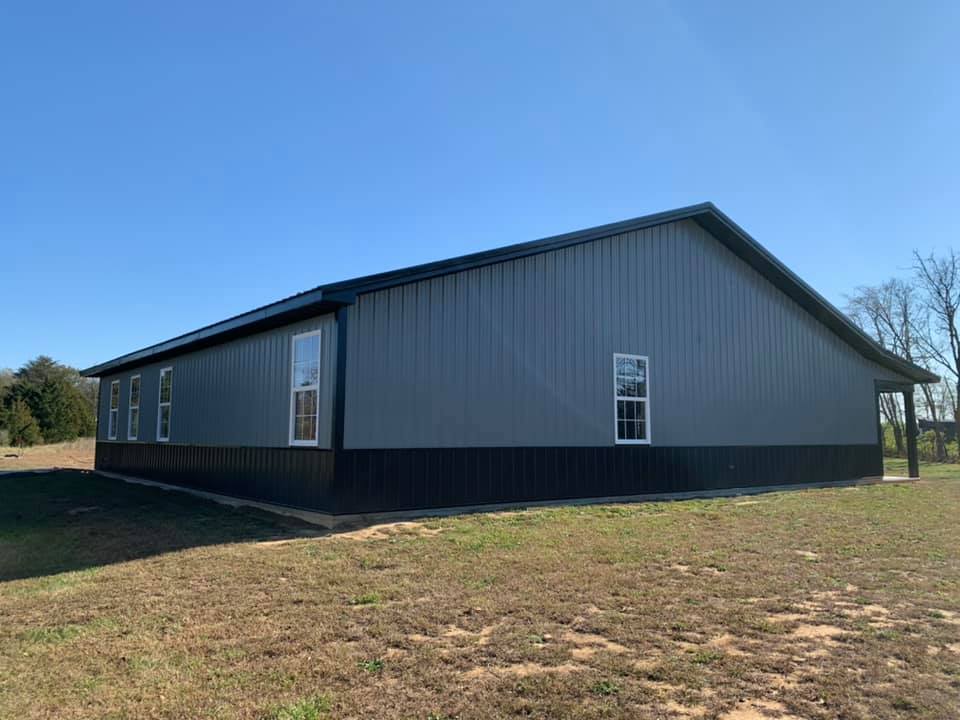 A metal-sided building with light gray walls and a black wainscot, featuring several windows, set in a grassy field.