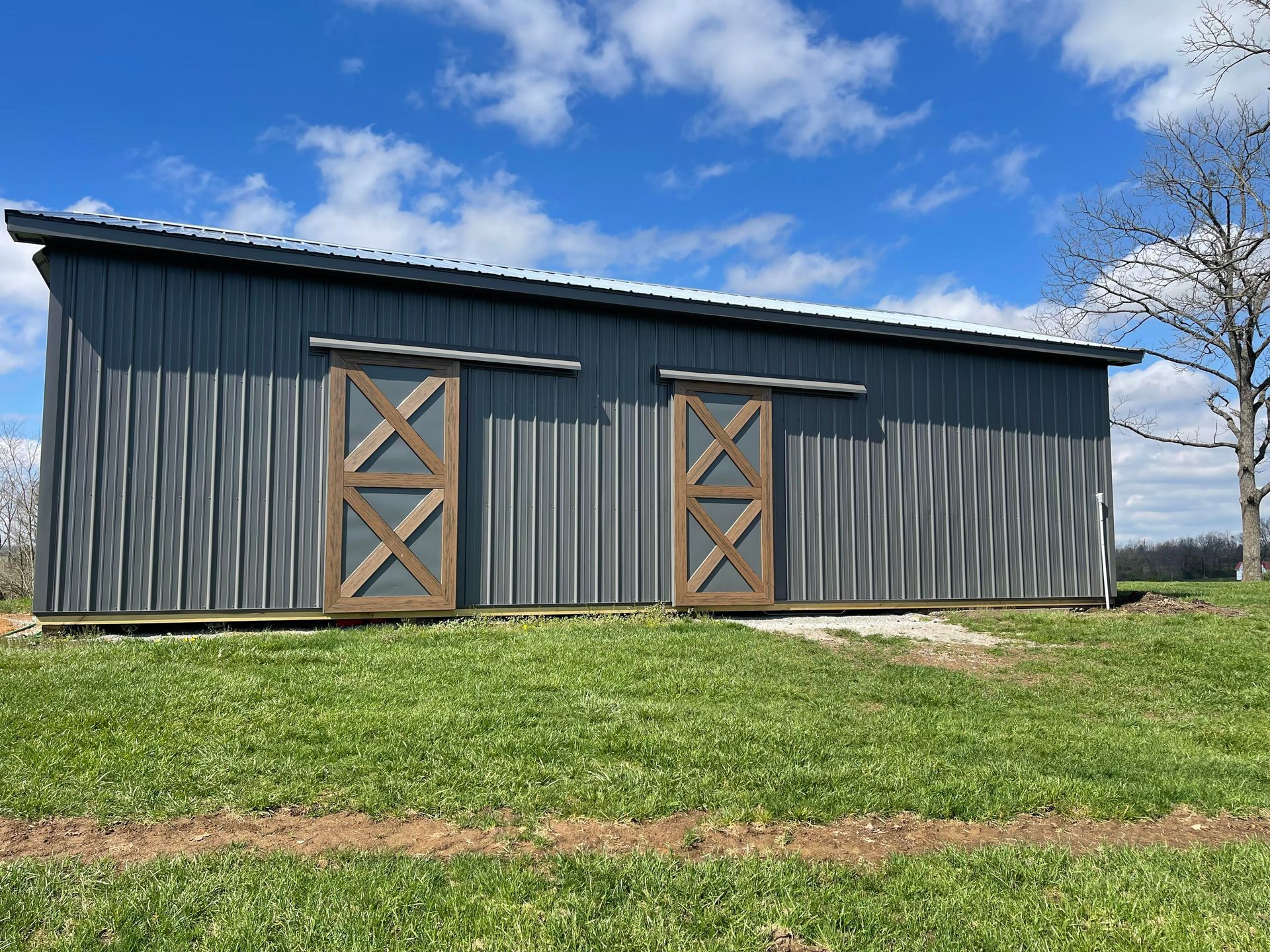 A gray metal barn with two brown sliding doors, set in a grassy field under a blue sky with scattered clouds.