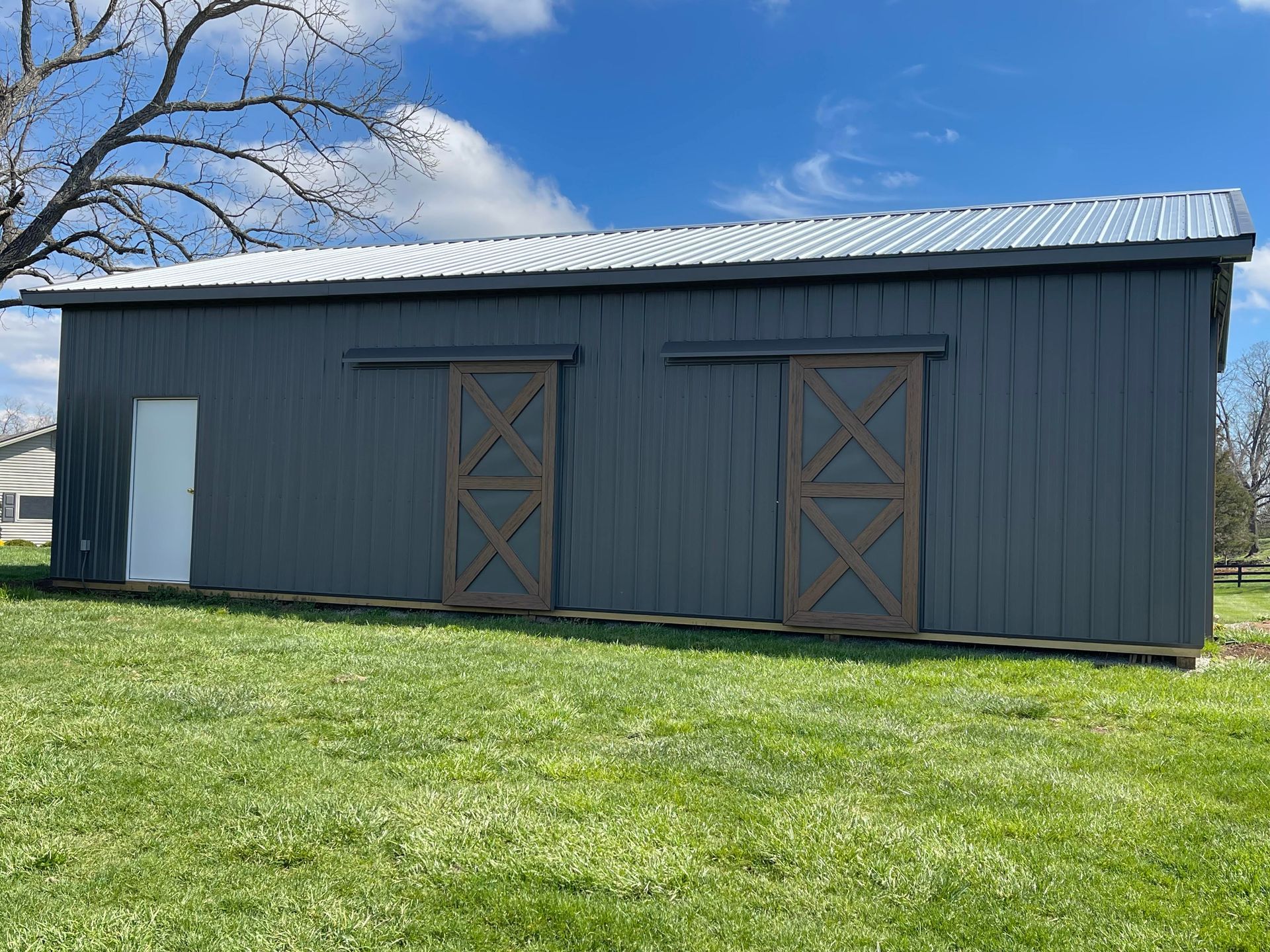 A dark gray barn with a white pedestrian door and two brown sliding barn doors, set in a grassy field under a blue sky.
