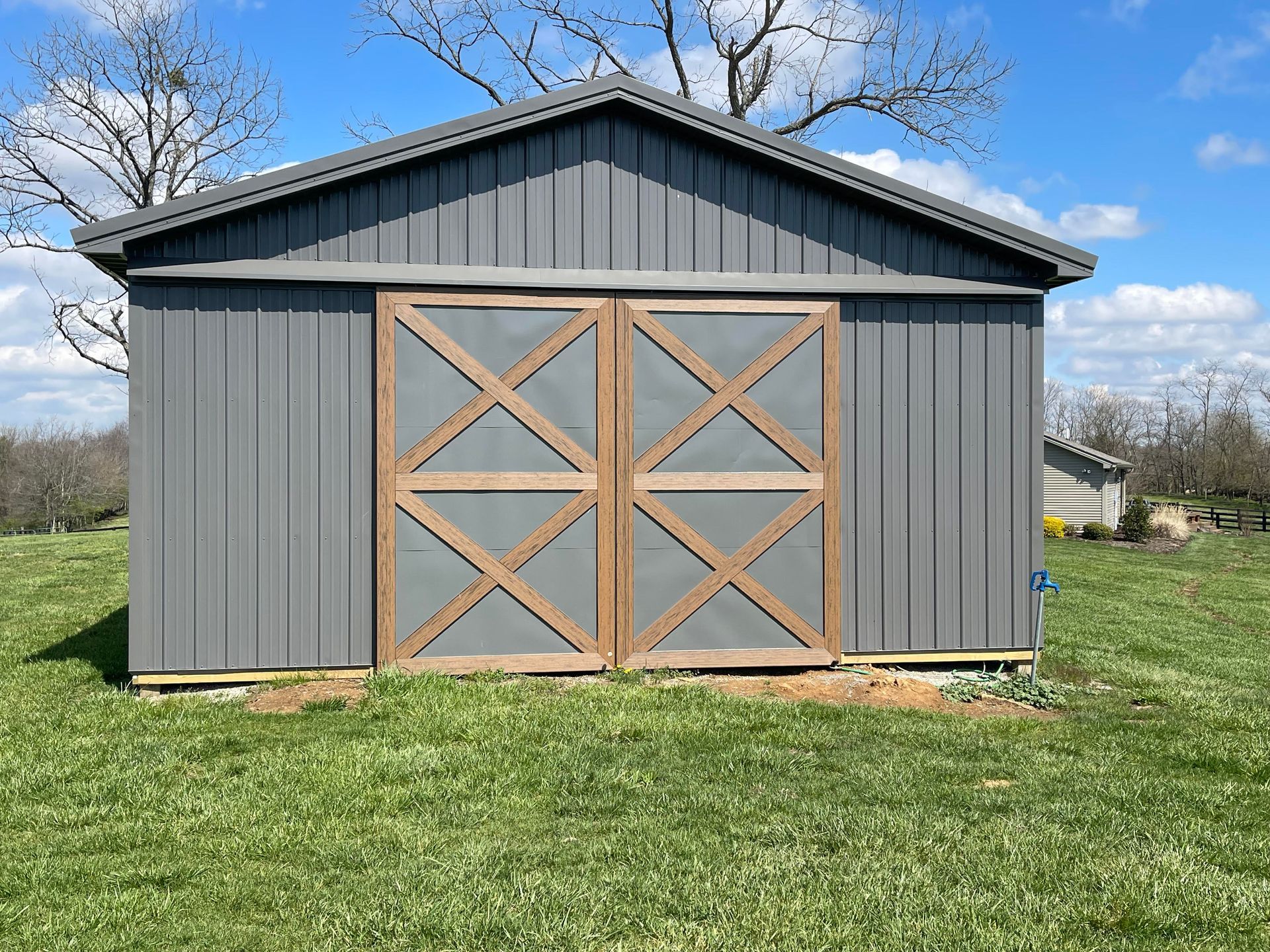 A gray metal shed with large double doors featuring brown cross-bracing, situated in a sunny grassy field.