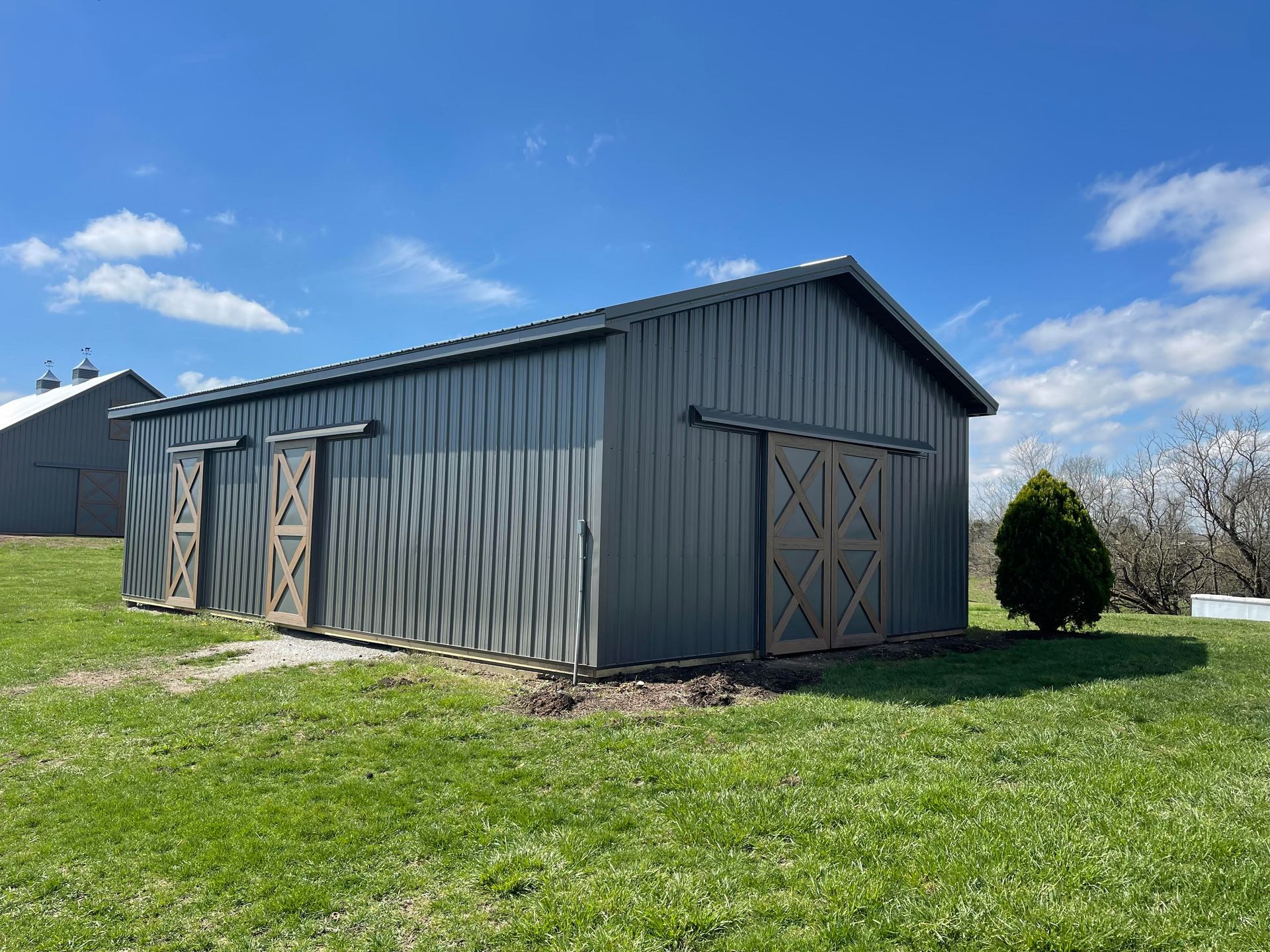A gray, metal-sided barn with brown sliding wooden doors, situated in a grassy field under a bright blue sky.