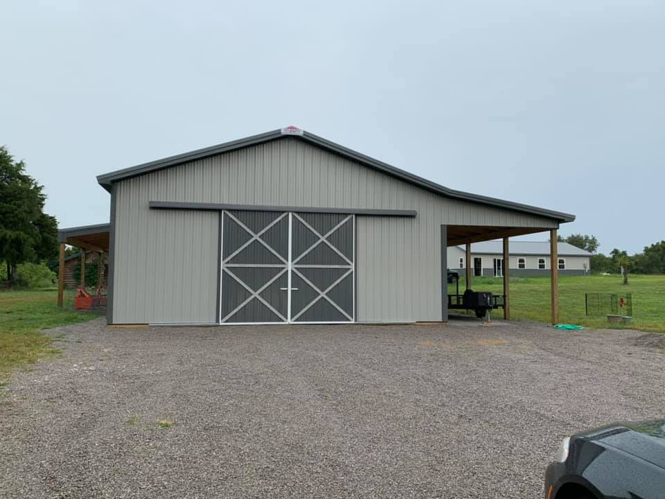 A grey metal barn with large sliding doors and open side lean-tos sits on a gravel lot under a cloudy sky.